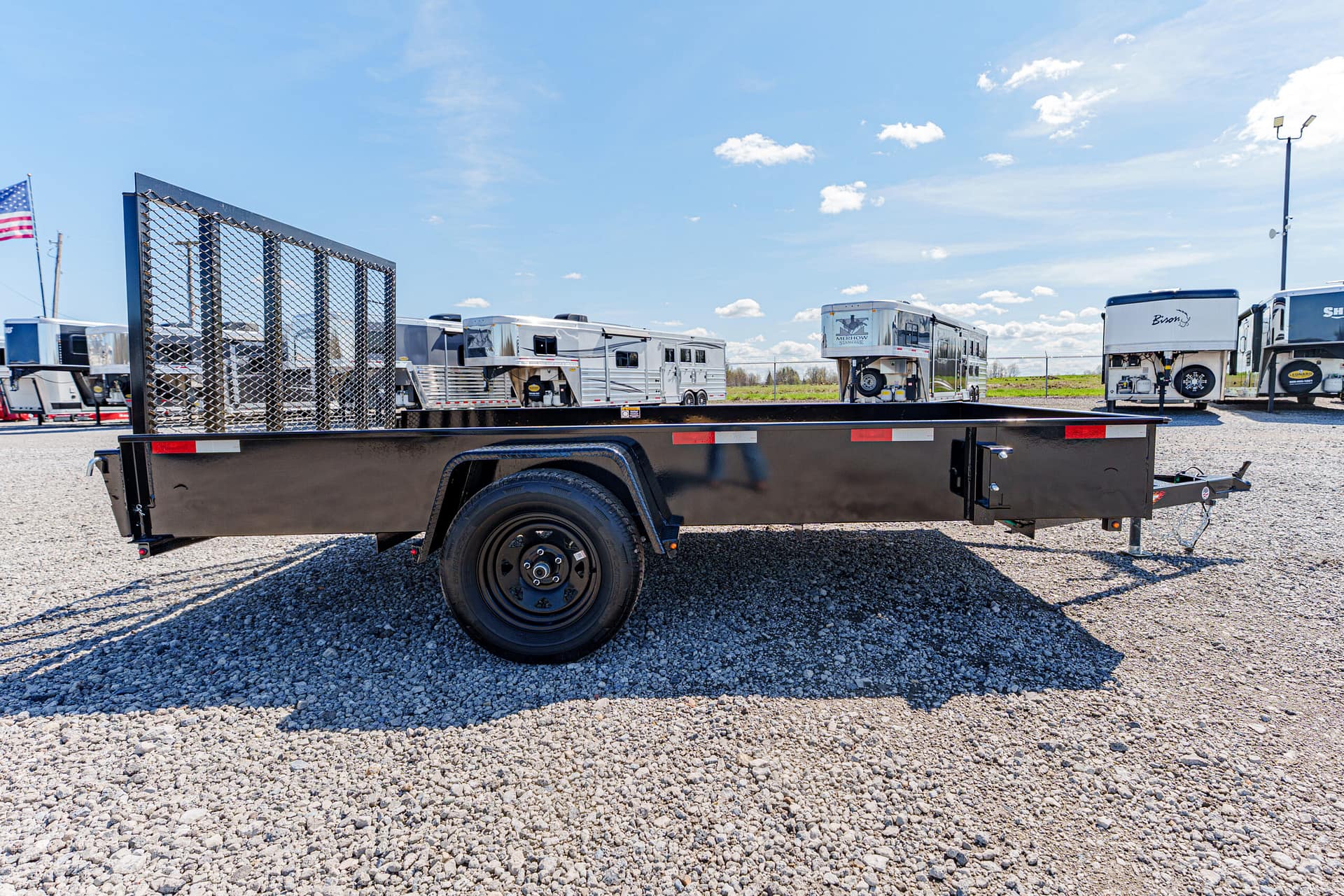 Black flatbed trailer with a metal mesh ramp in a gravel lot, with horse trailers in the background under a blue sky.