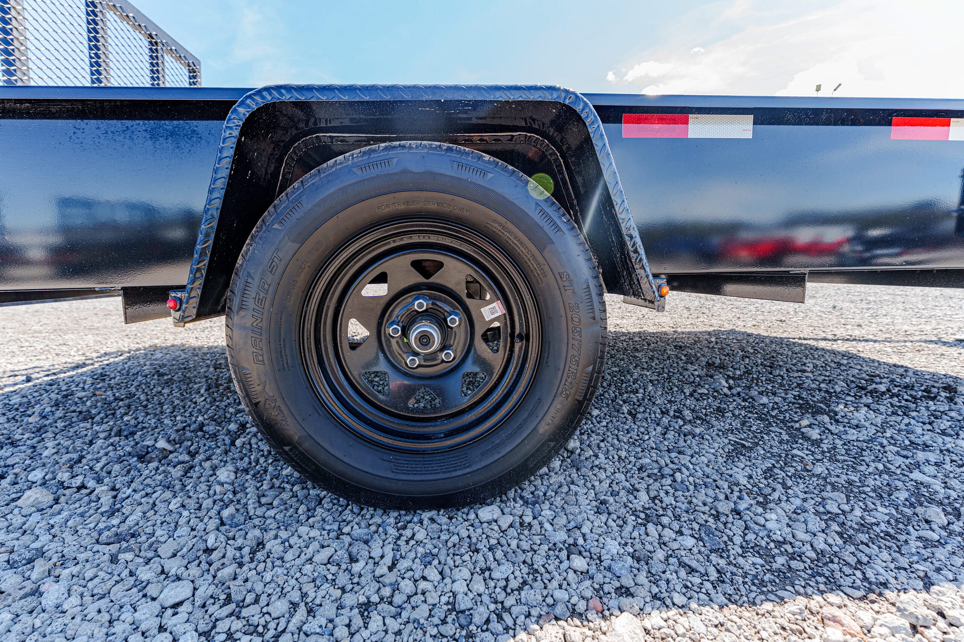 Close-up of a black trailer wheel and curved metal fender on a blue trailer, with gravel ground and a clear sky above.