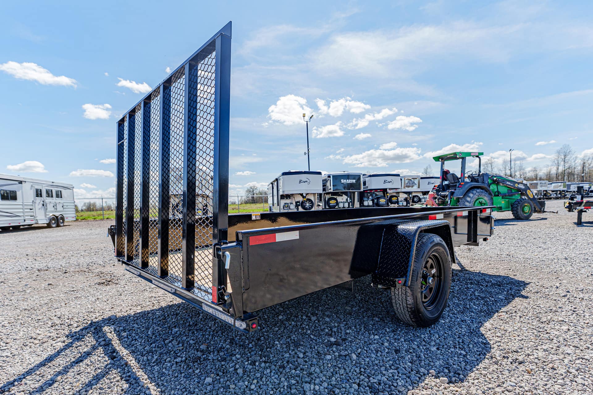Black utility trailer with a tall mesh ramp gate on a gravel lot, under a blue sky with distant farm equipment visible in the background.