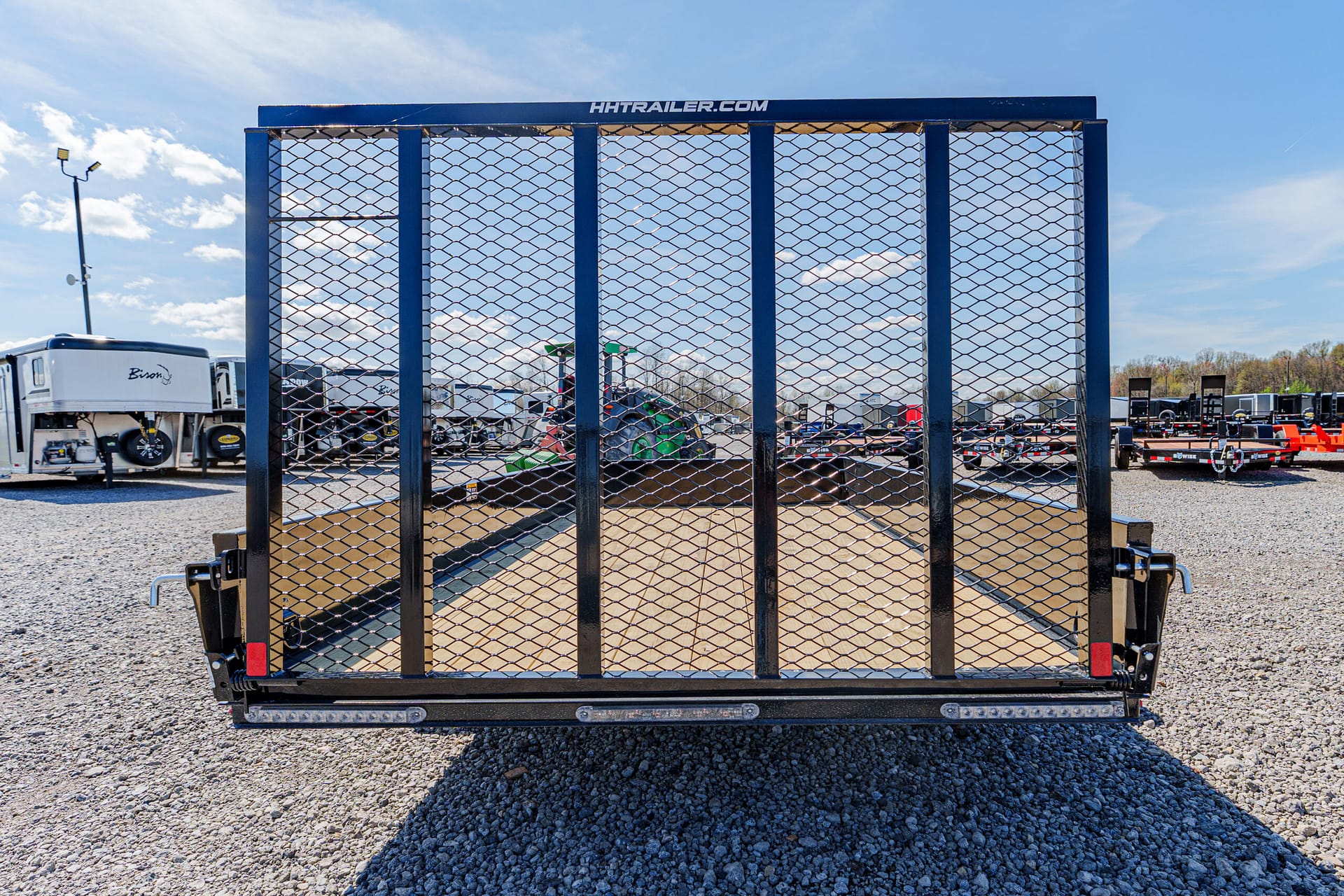 Front-on view of a blue utility trailer with a diamond-mesh gate in a gravel lot; 'HTTRAILER.COM' label along the top edge.