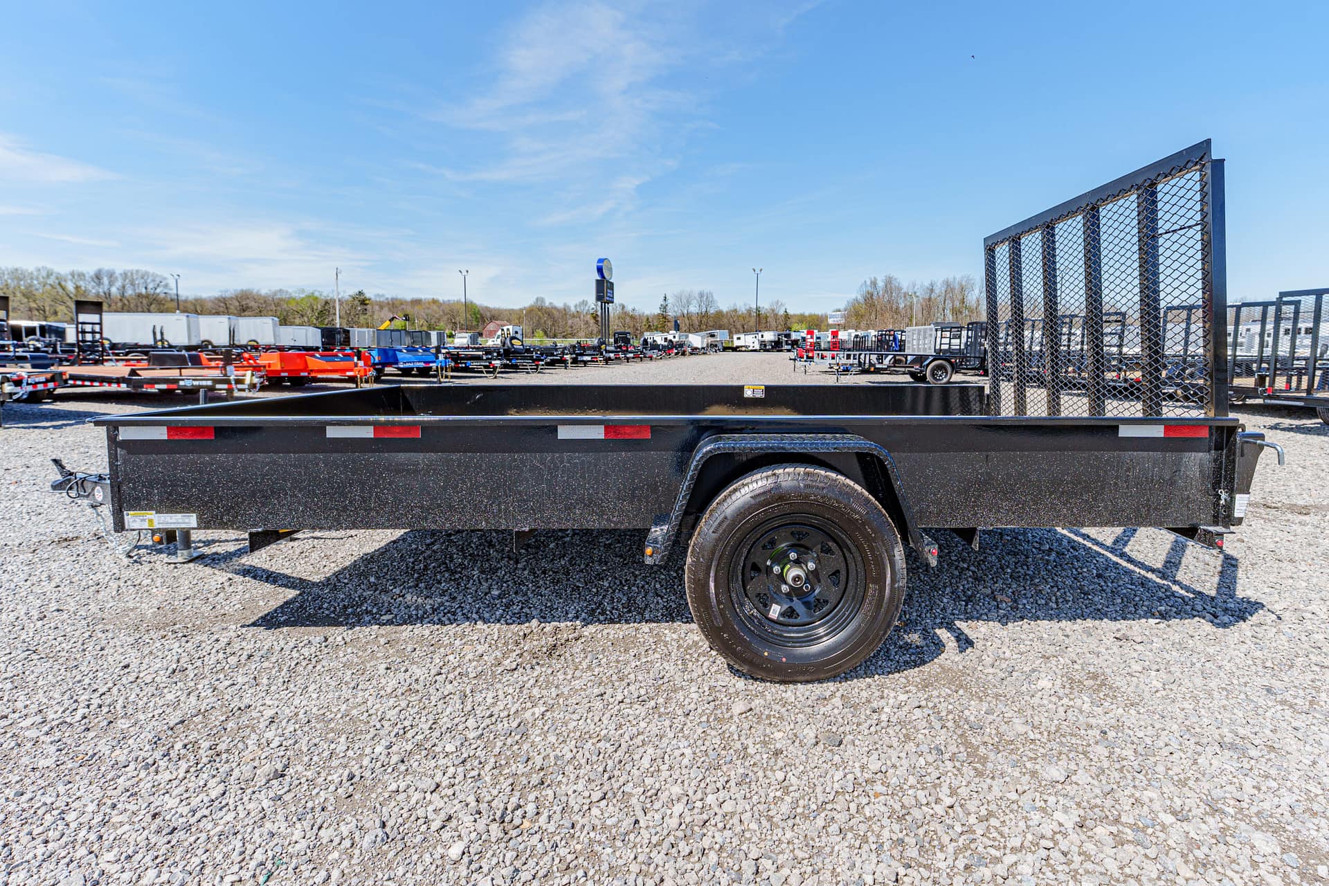 Black flatbed trailer with a tall mesh divider in a gravel lot, with numerous trailers lined up in the background.
