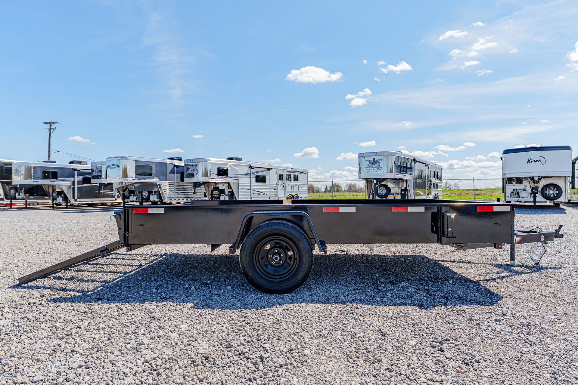 Line of travel trailers and a flatbed trailer parked on a gravel lot under a blue sky with scattered clouds.