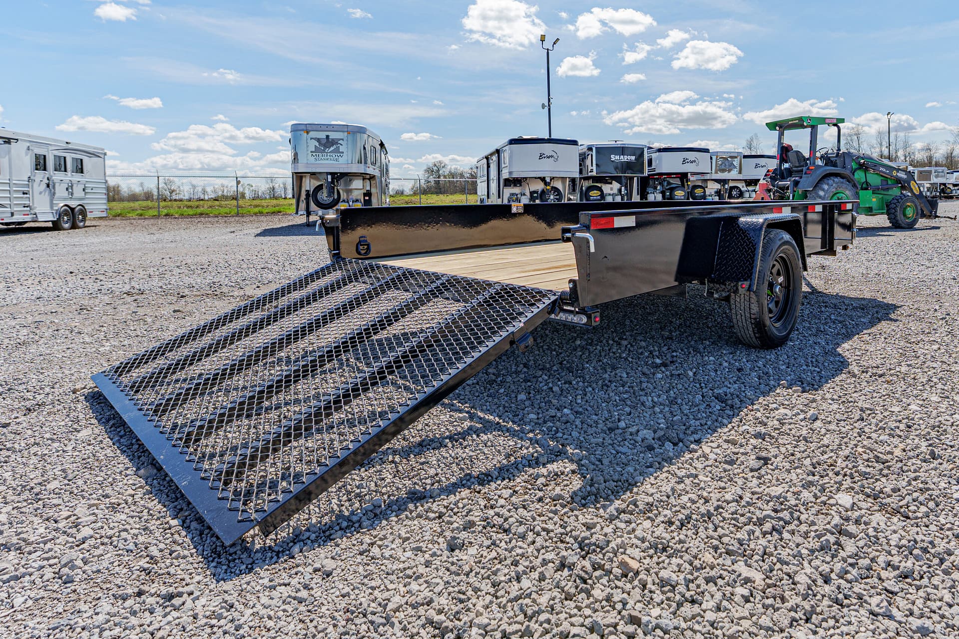 Flatbed trailer with a lowered mesh ramp on a gravel lot, with horse trailers and a green tractor in the background under a blue sky.