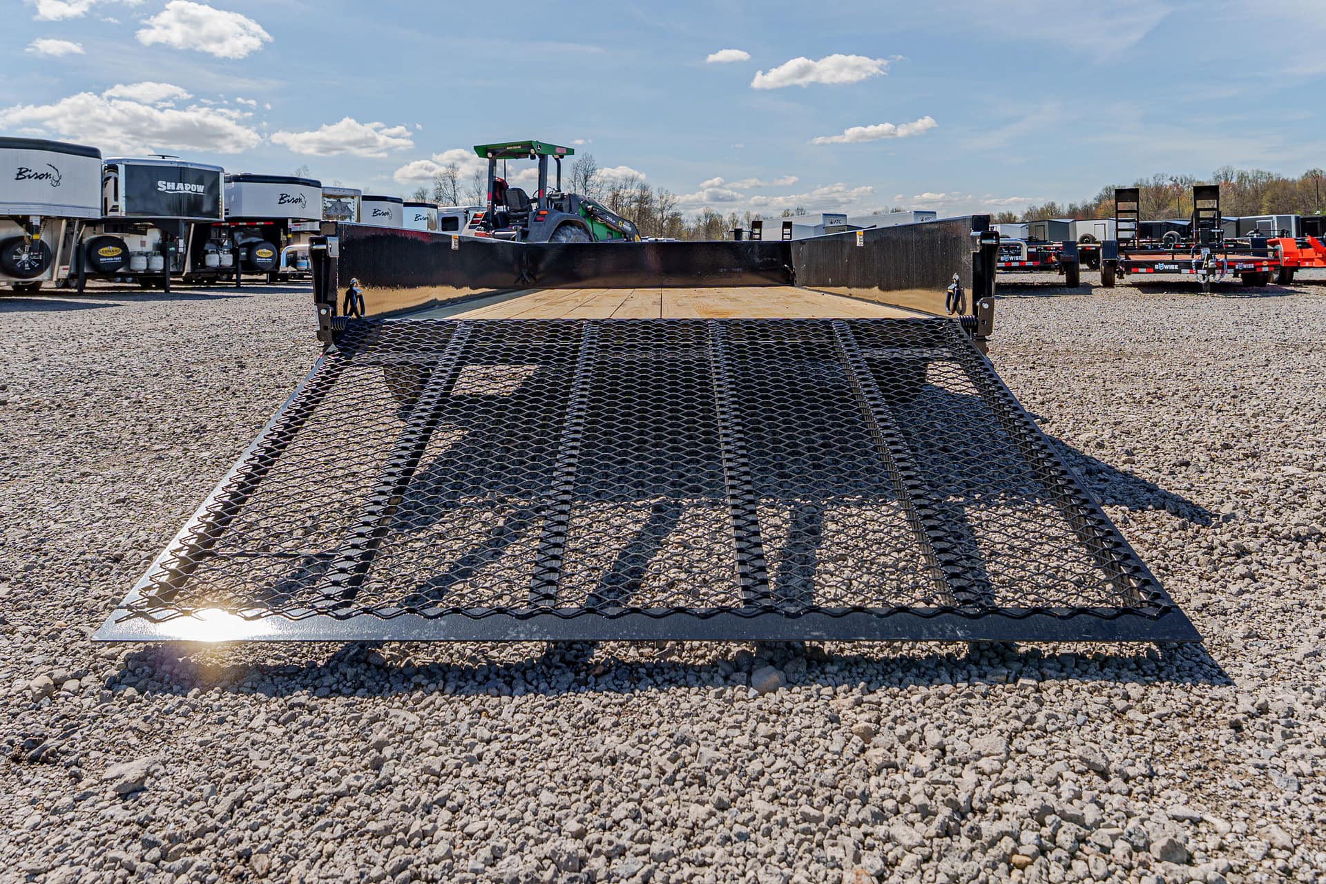 Wide shot of a black metal grate ramp on a flatbed trailer in a gravel lot, with horse trailers and a green tractor in the background under a blue Sky