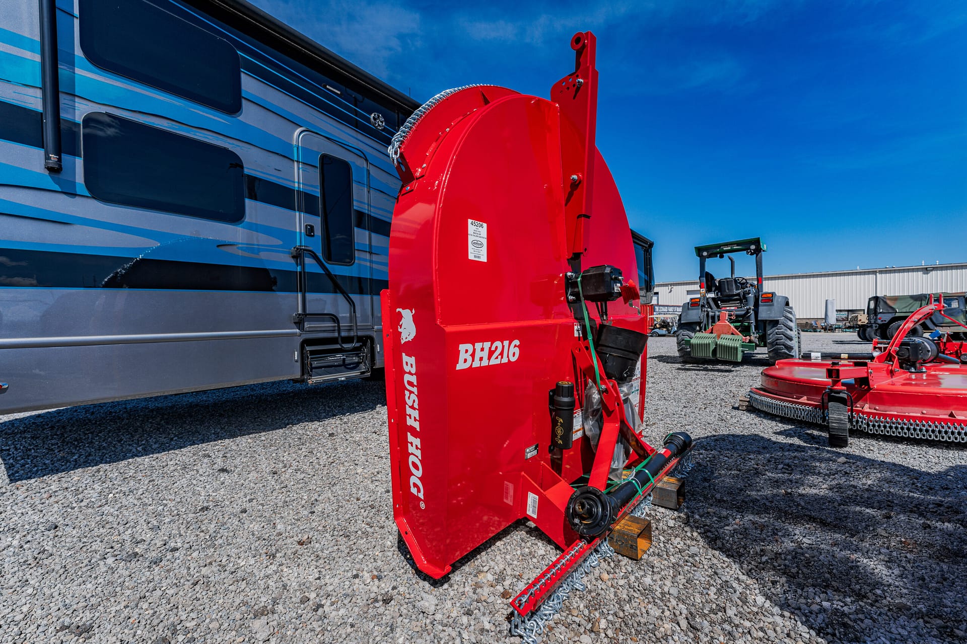 Red Bush Hog BH216 rotary mower attachment on gravel beside a blue RV, with tractors in the background (outdoor equipment yard).