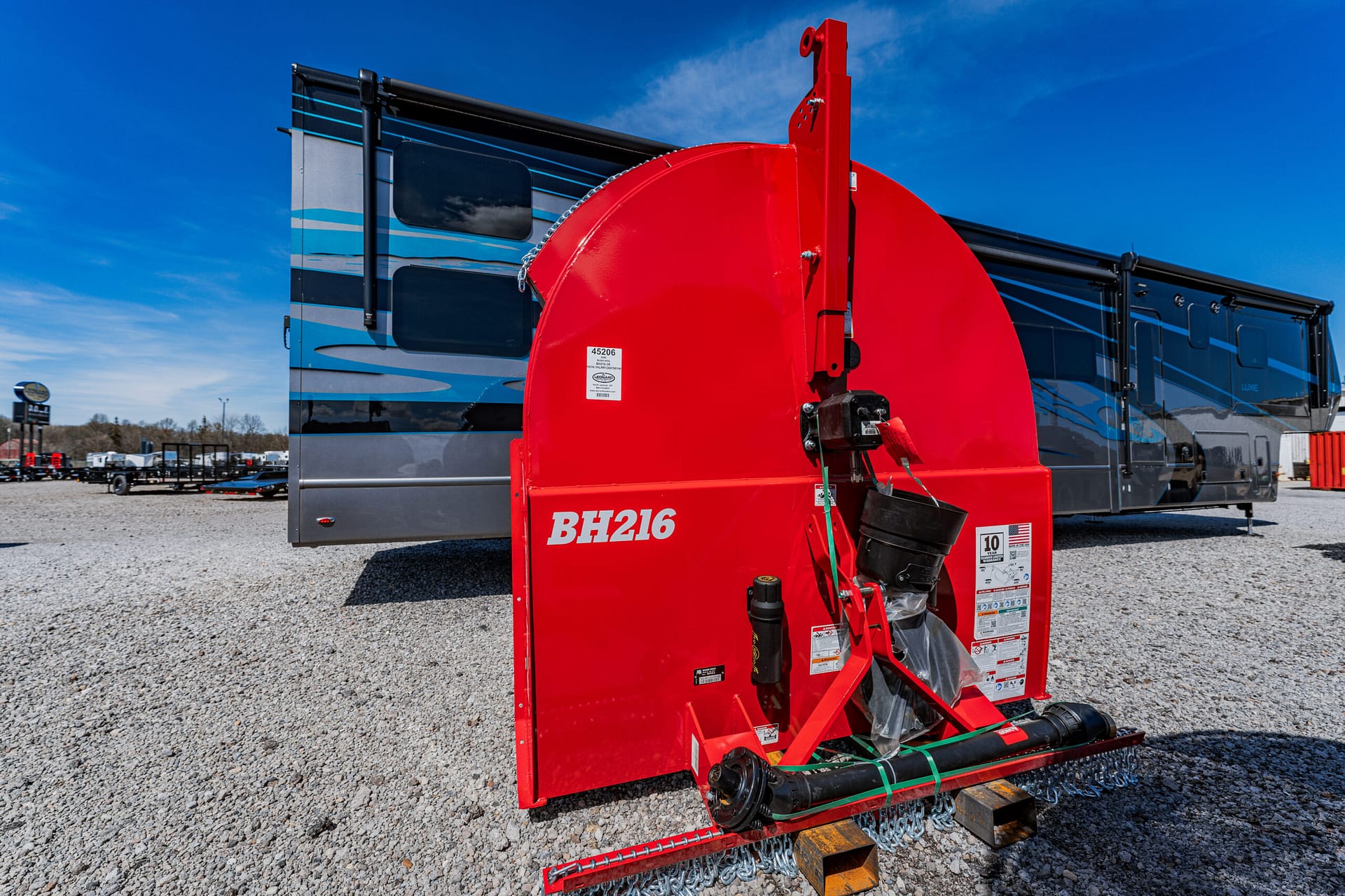 Red BH216 wood chipper attachment mounted on a trailer in a gravel lot, with a blue RV in the background.