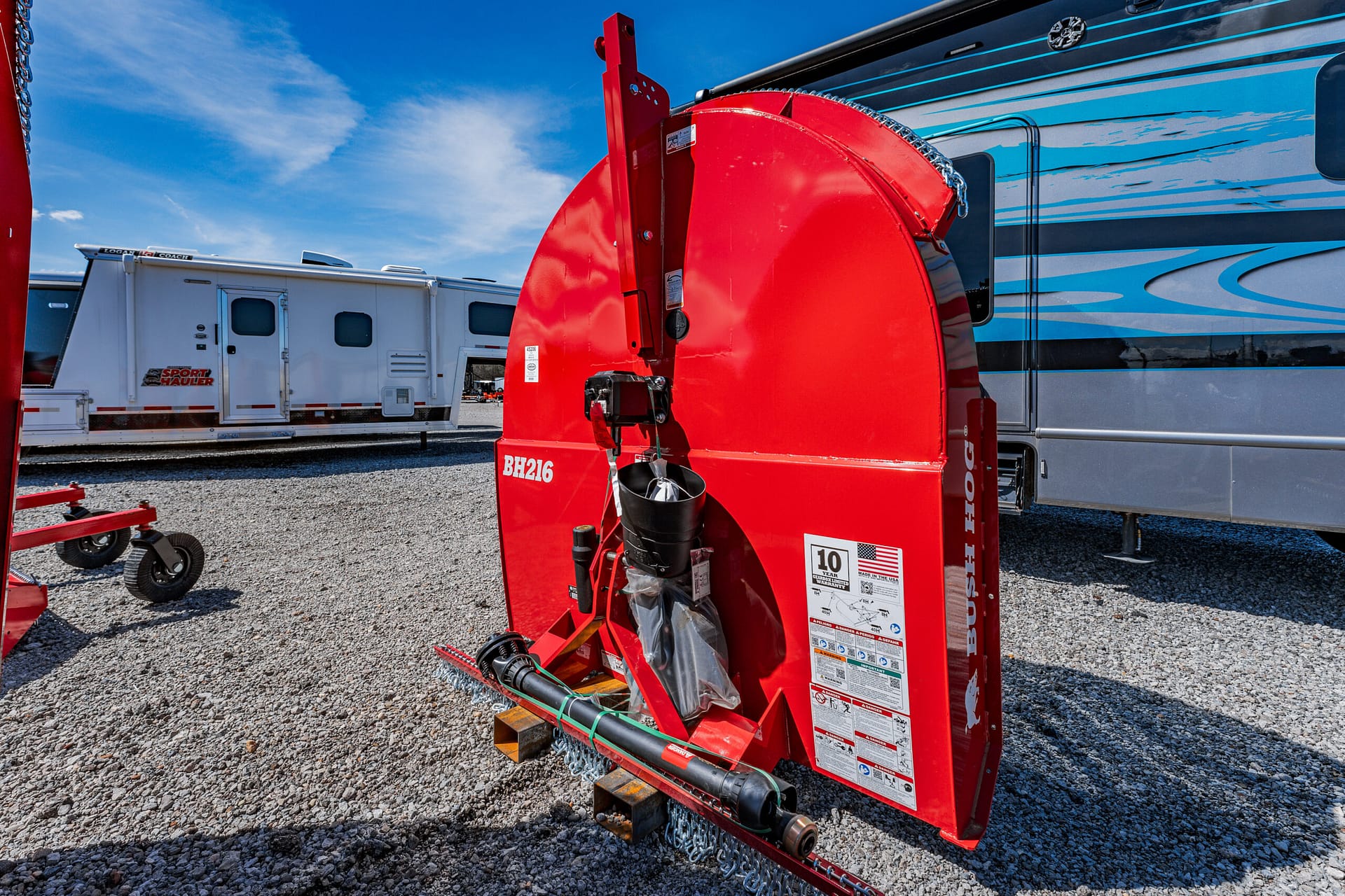 Red circular industrial snow blower attachment on a trailer frame, with a black driveshaft and warning labels, resting on gravel beside an RV park setup.