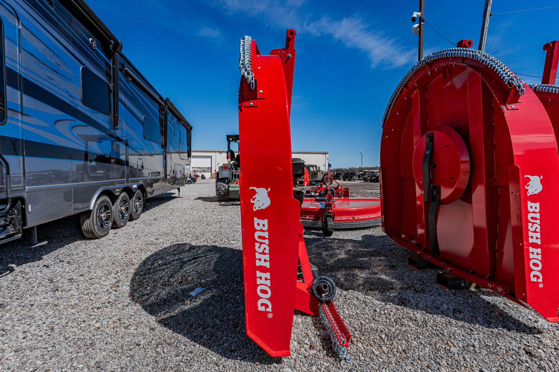 Red Bush Hog rotary cutter components on a gravel lot with a blue RV trailer to the left and a clear blue sky above.