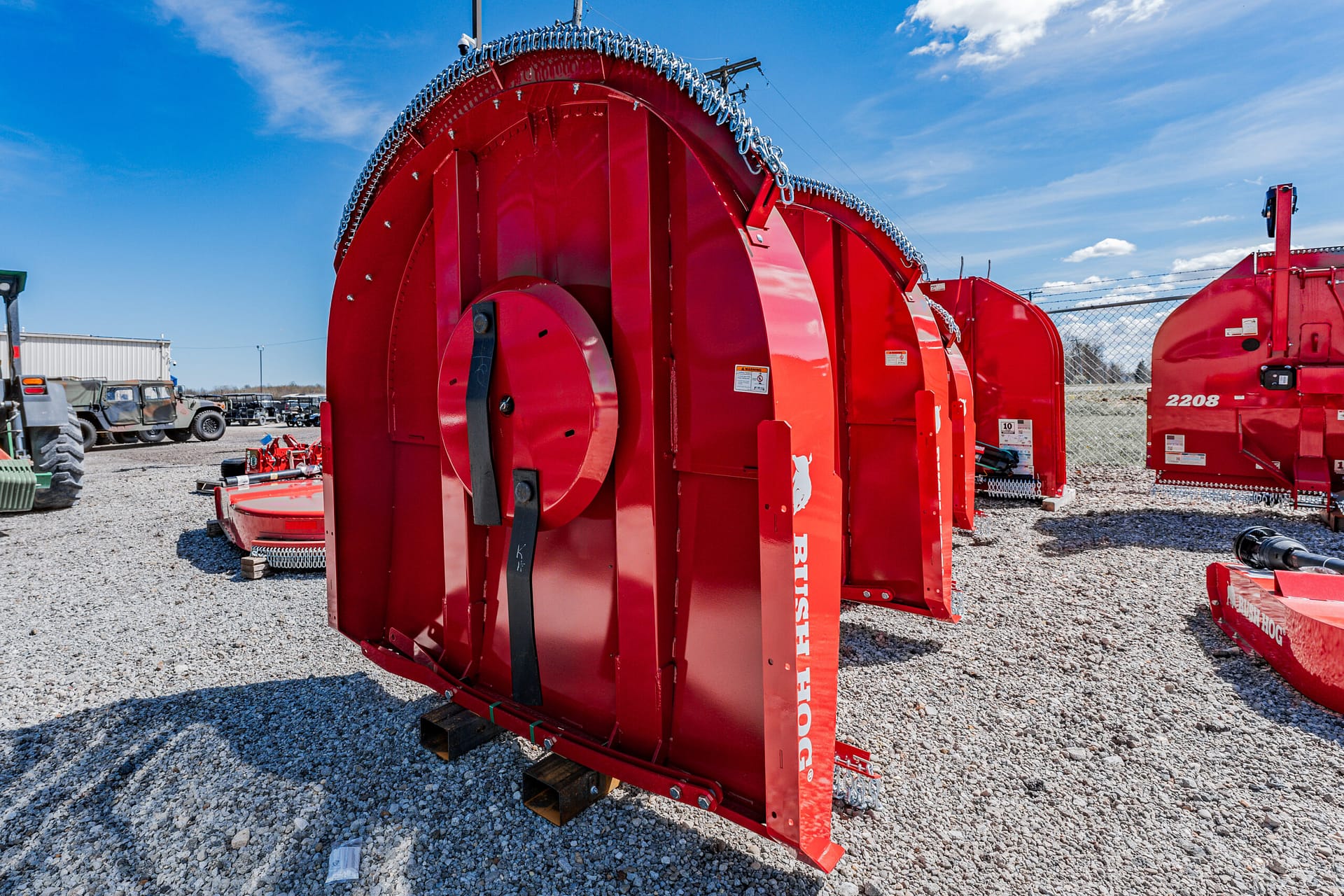Large red circular agricultural machine housing with chain guard on a gravel lot, surrounded by other red farm equipment.