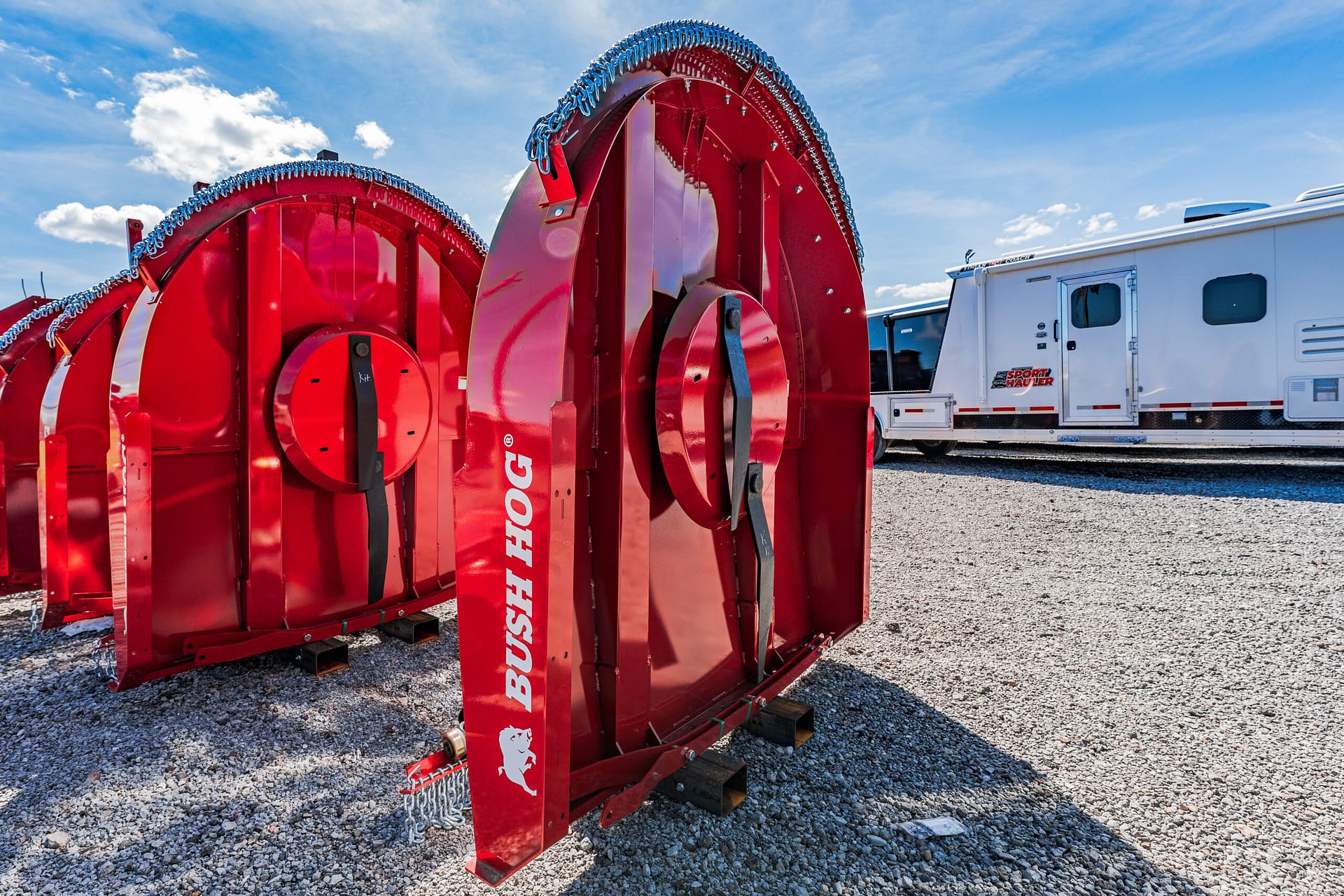 Two large red Bush Hog rotary mower sections sit on a gravel lot, with a white RV/trailer in the background.
