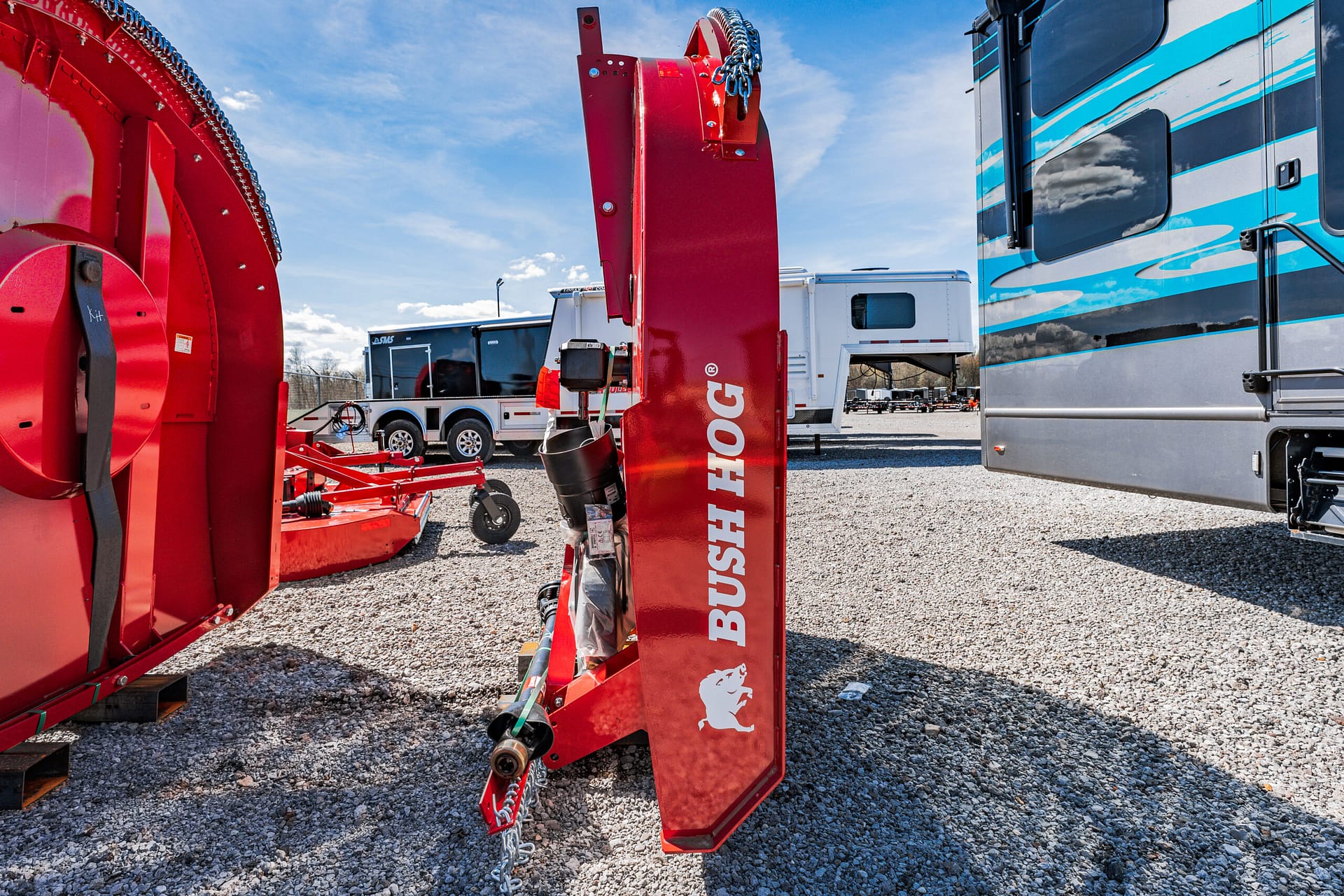 Red Bush Hog rotary cutter standing upright in a gravel lot with RVs and another red mower in the background.