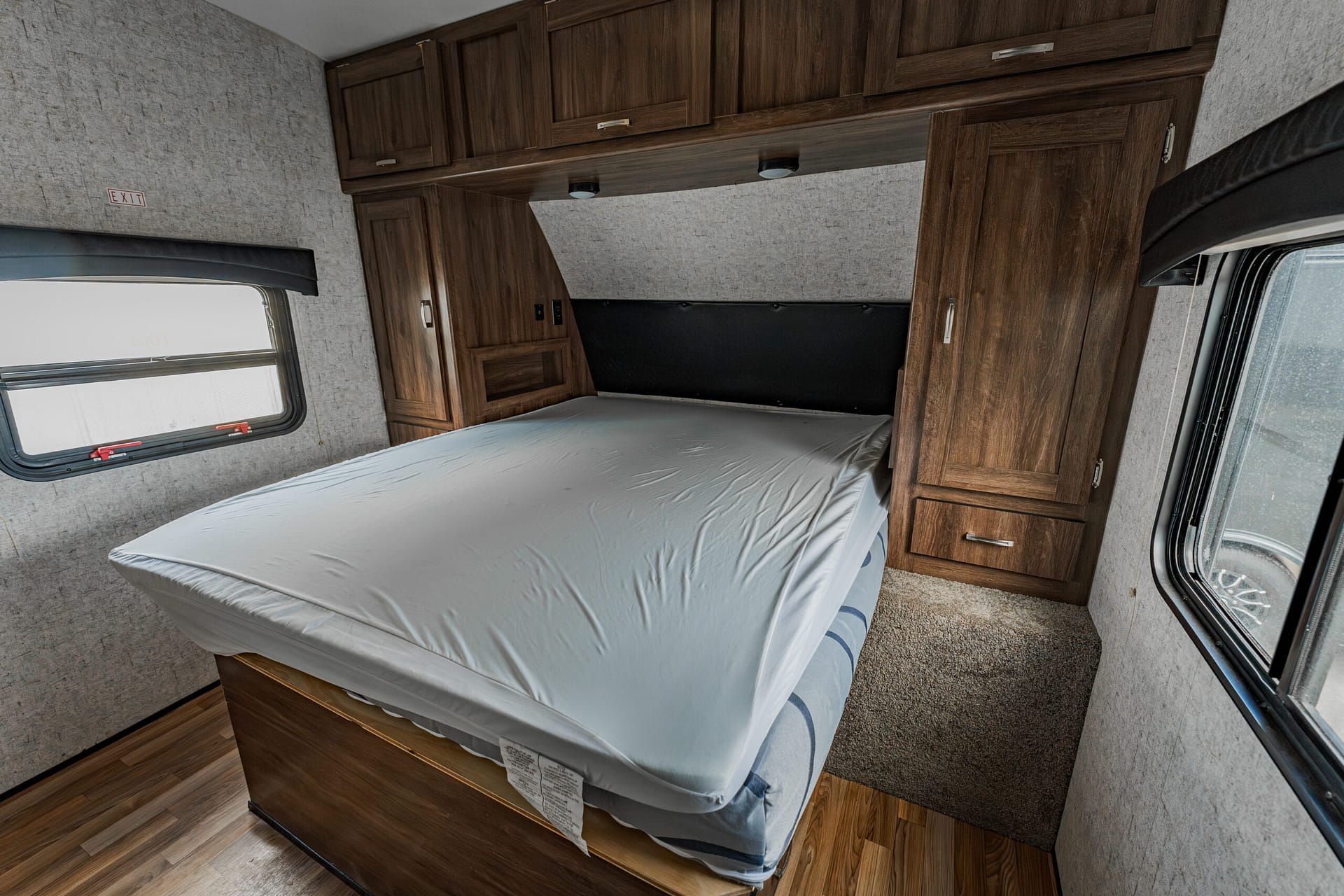 Bedroom inside a camper or RV with a full bed, dark wood cabinets above and around, and a window with a shade on the left side