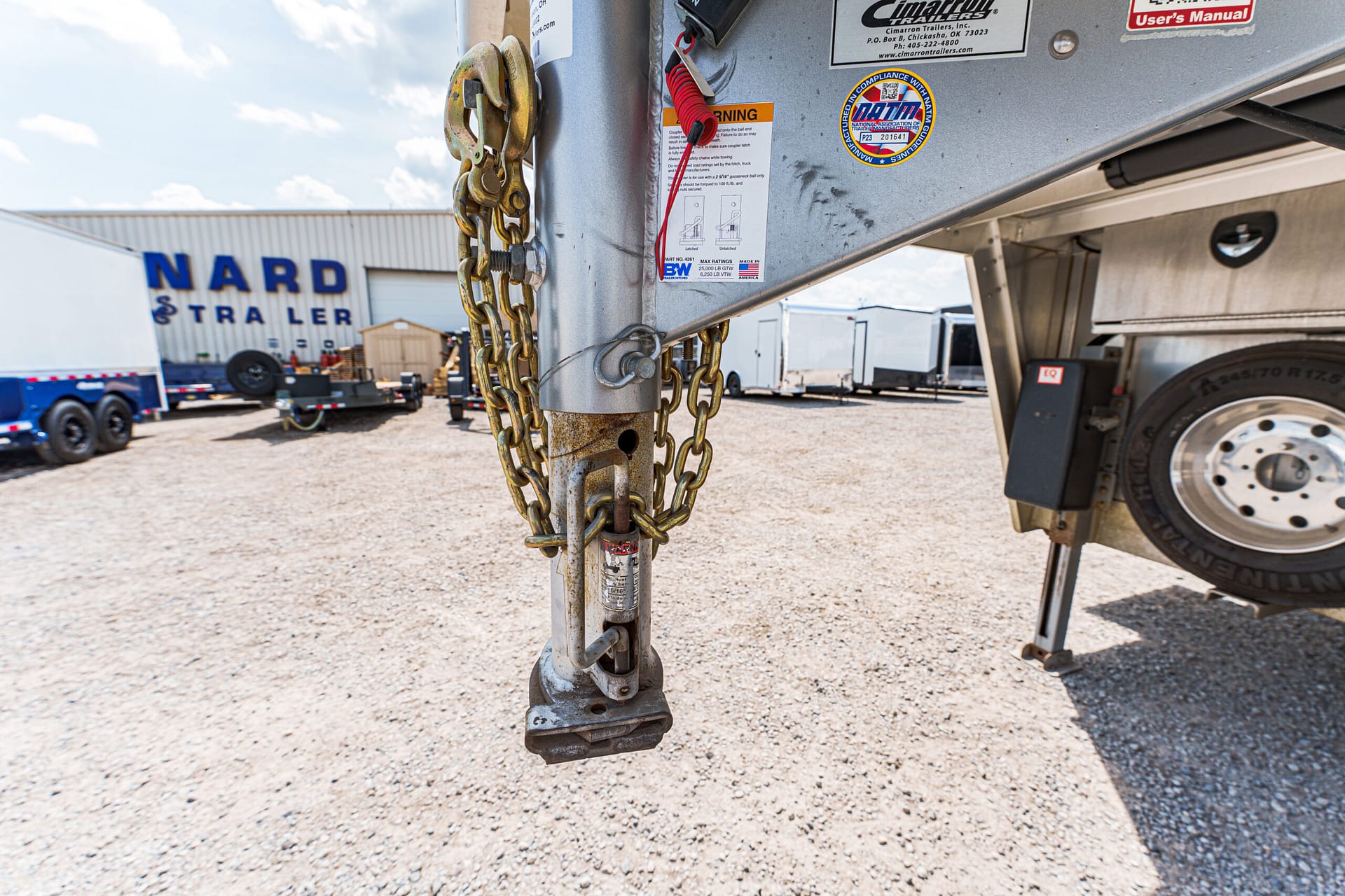 Close-up of a rusted trailer hitch with safety chains and a pin in a gravel yard, other trailers in the background.