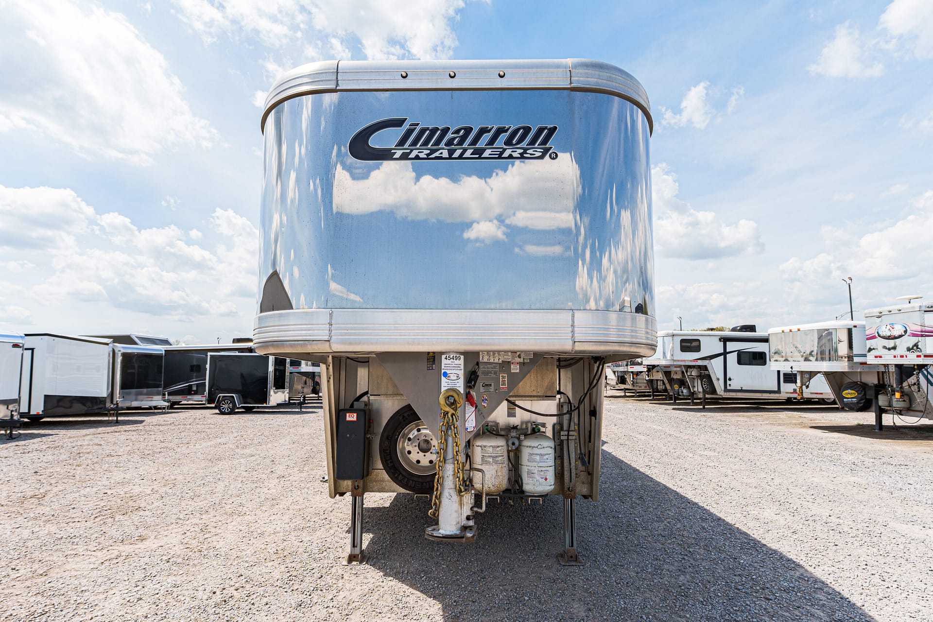 Front view of a polished Cimarron gooseneck trailer in a gravel lot with other trailers and a blue sky behind it, logo visible on the front