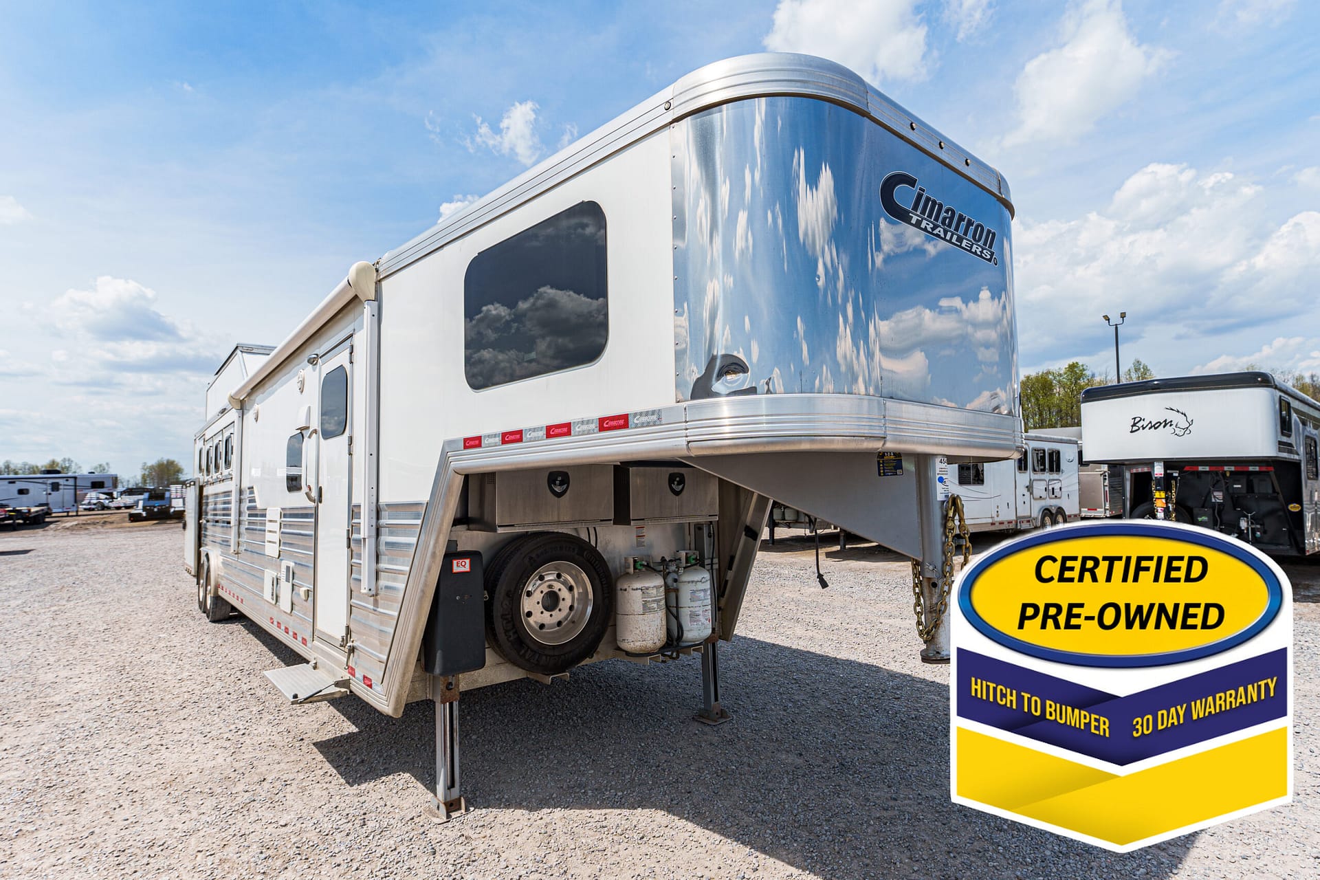 Shiny silver gooseneck trailer with a Cimarron Trailers logo, parked on a gravel lot beside a large yellow 'Certified Pre-Owned' sign under a blue sky.