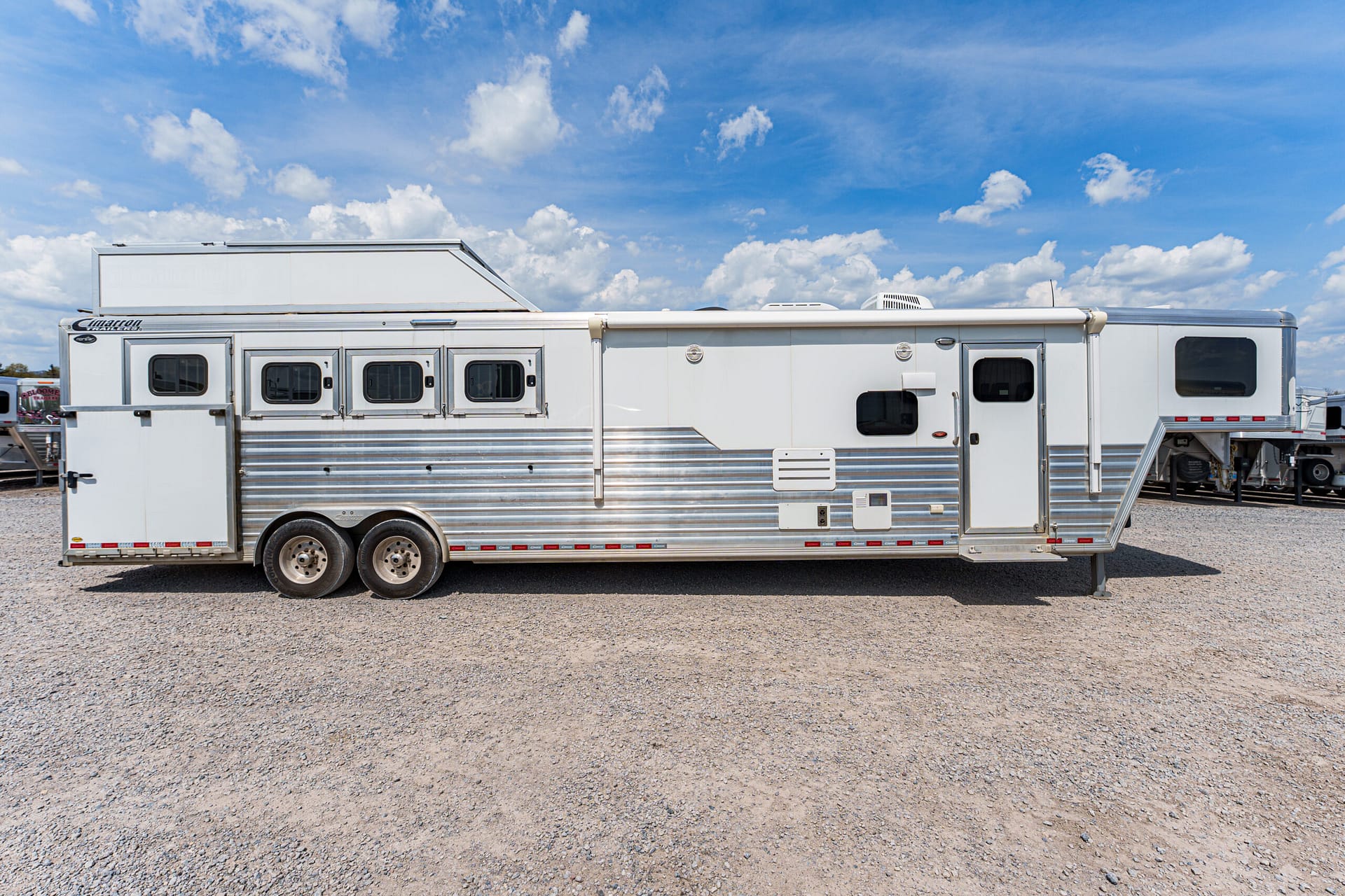 Long white horse trailer with aluminum siding and several windows, parked on a gravel lot under a blue sky with scattered clouds
