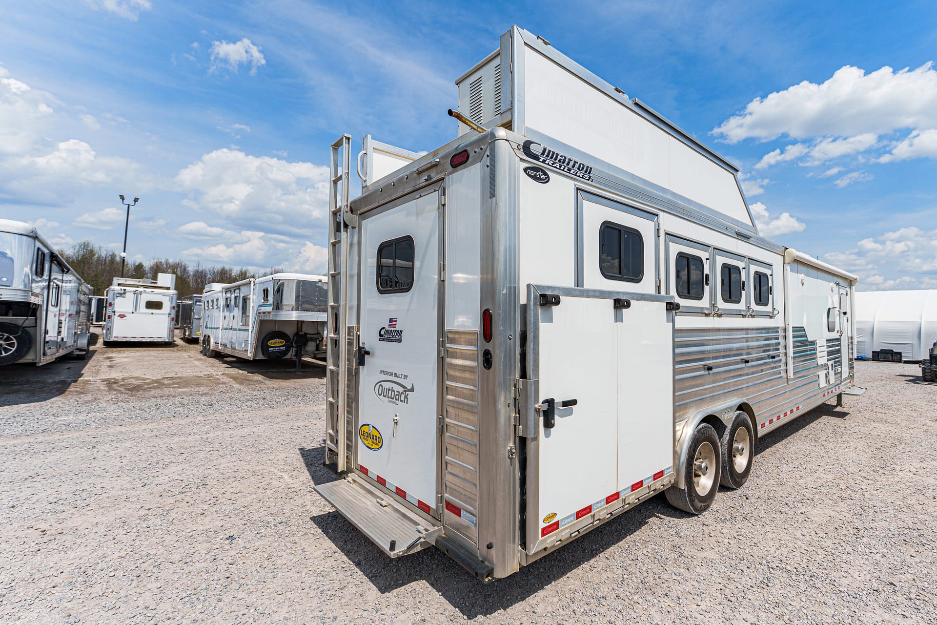Silver horse trailer in the foreground on a gravel lot with other trailers lined up under a bright blue sky with clouds.