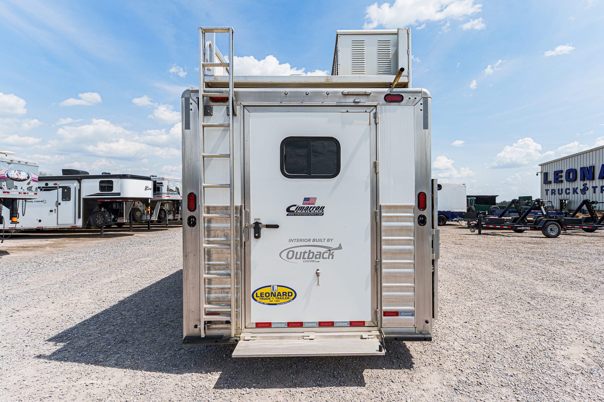 Rear view of a silver aluminum trailer with a vertical ladder on the left, parked in a gravel lot with other trailers under a blue sky.