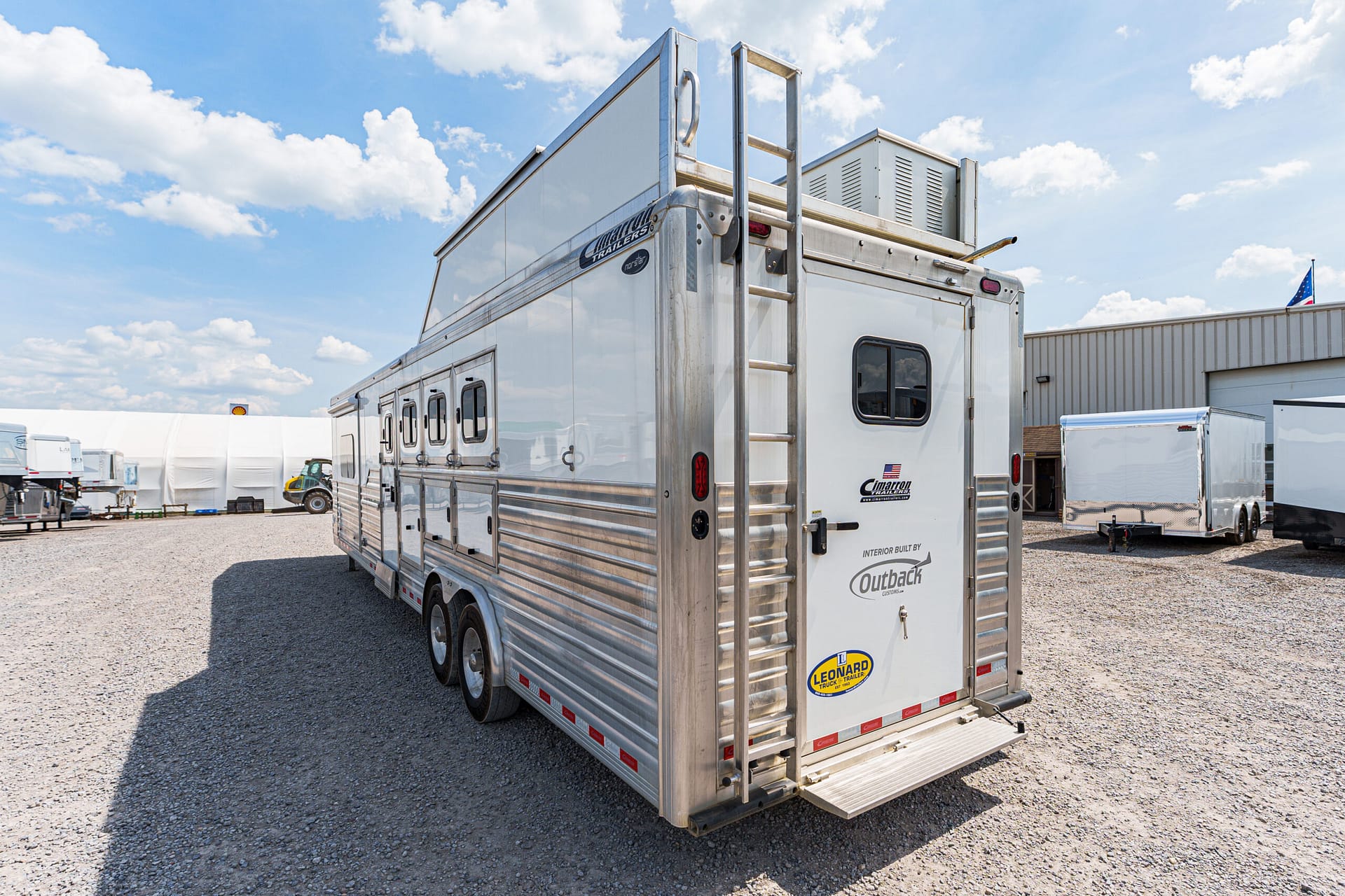 Silver aluminum horse trailer with multiple stalls and a rear ladder, parked on a gravel lot beneath a blue sky with clouds.