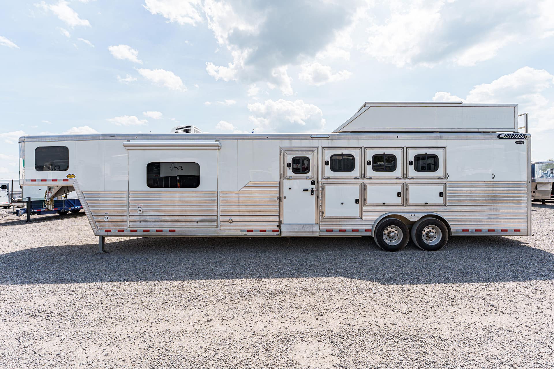 Long silver horse trailer parked on gravel with multiple doors and windows under a blue sky with clouds