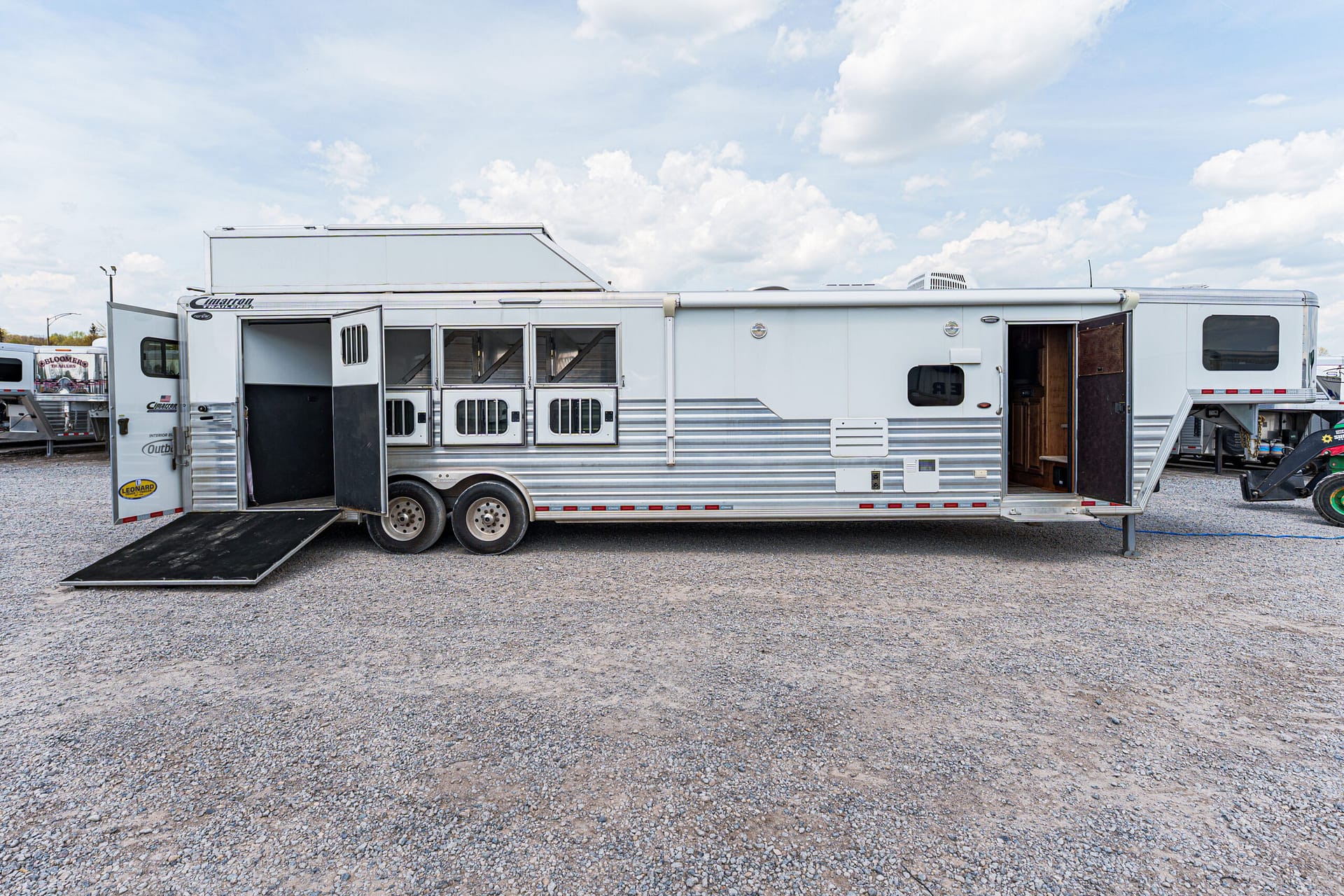 Silver horse trailer with an extended ramp down on a gravel lot and open rear doors, ready for loading horses under a partly cloudy sky.
