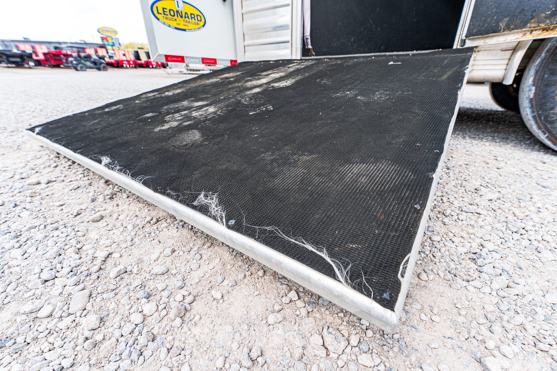 Black metal trailer loading ramp on a gravel lot, with scuffs and dirt marks visible. Behind it are other trucks and trailers.