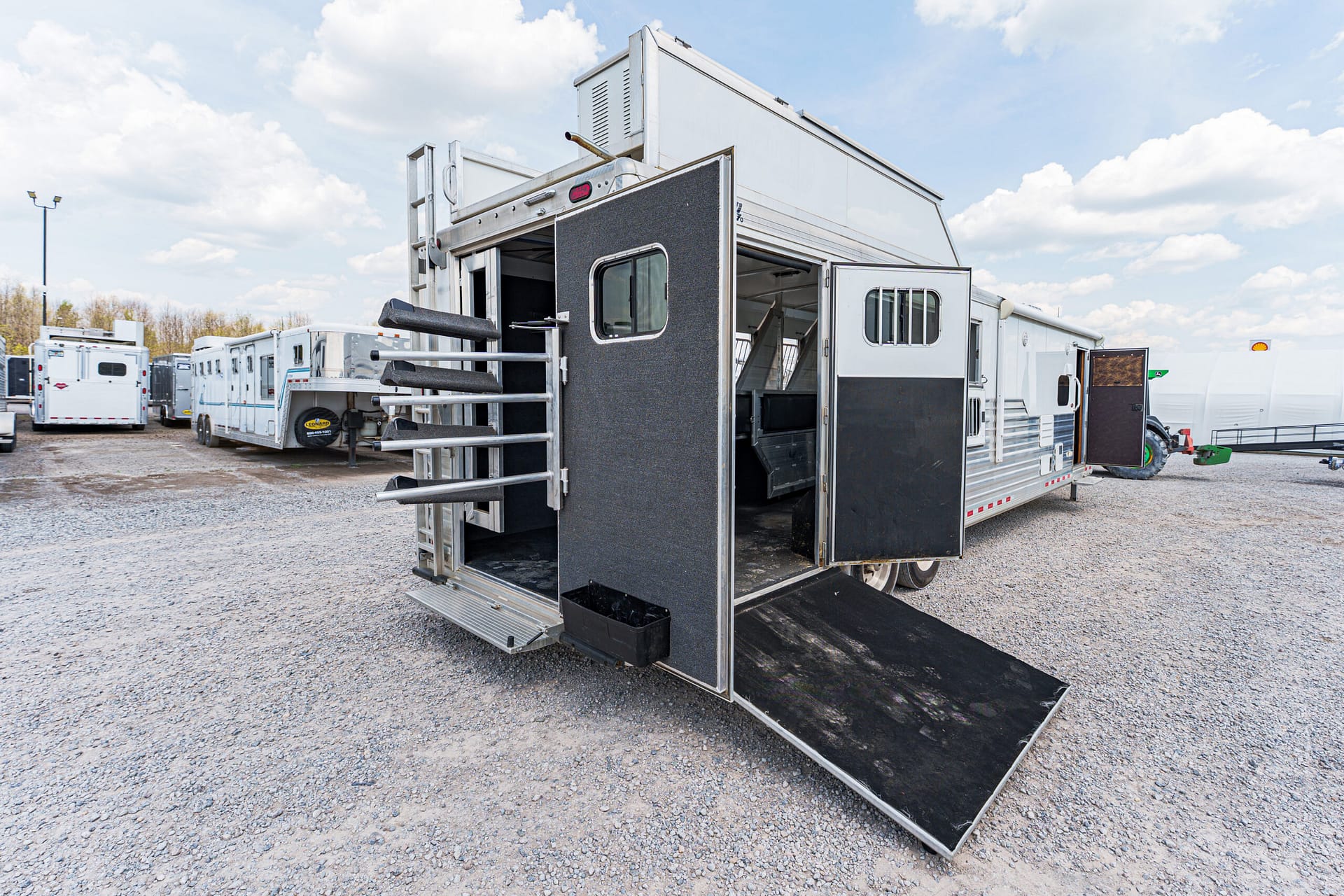 Silver horse trailer with an open ramp, parked among other trailers on a gravel lot under a cloudy sky.