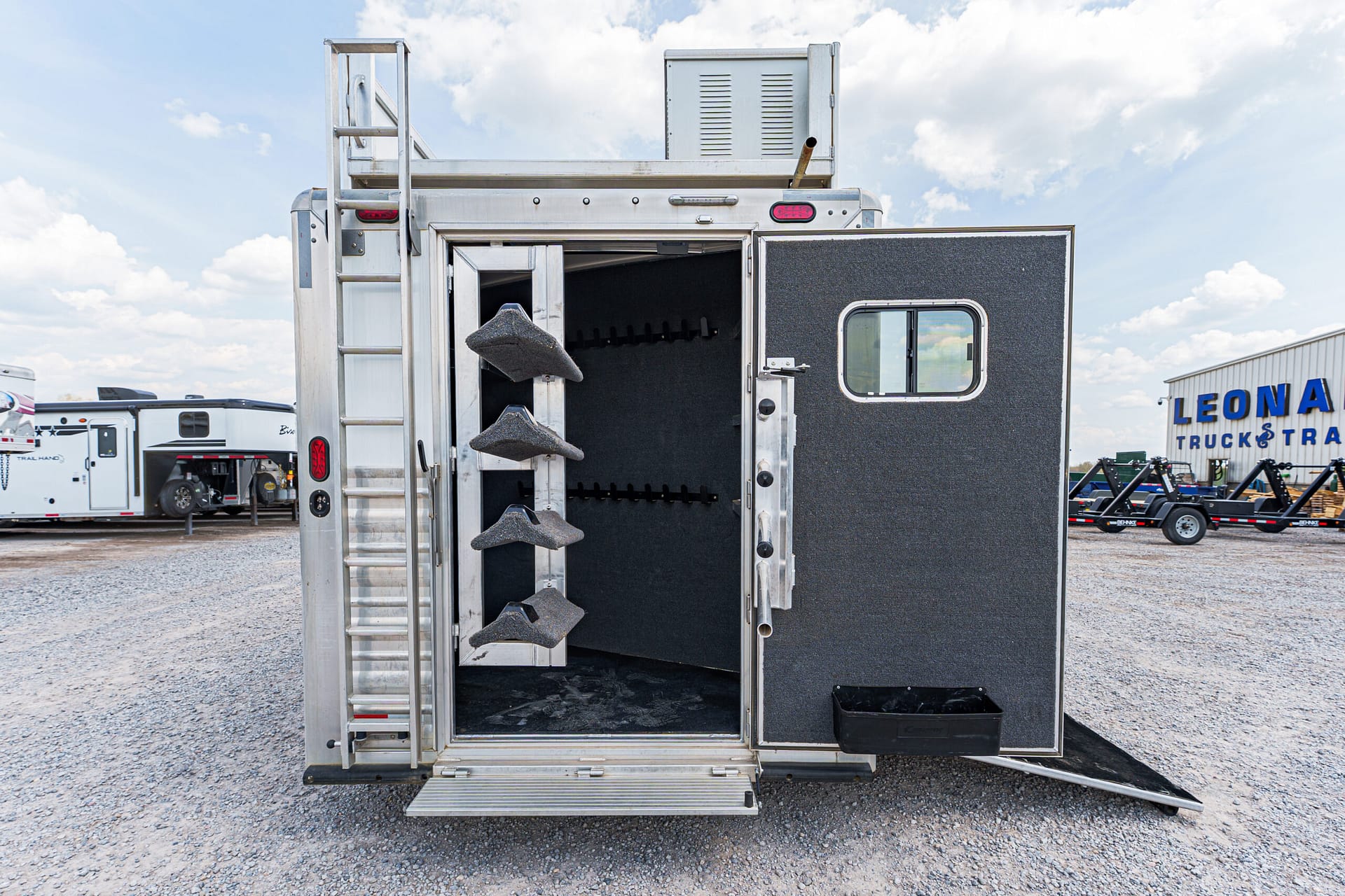 Rear of a metal trailer with open back door, showing stairs and a ramp, in a gravel lot at a truck trailer sales yard with other trailers in the background and a blue sky.