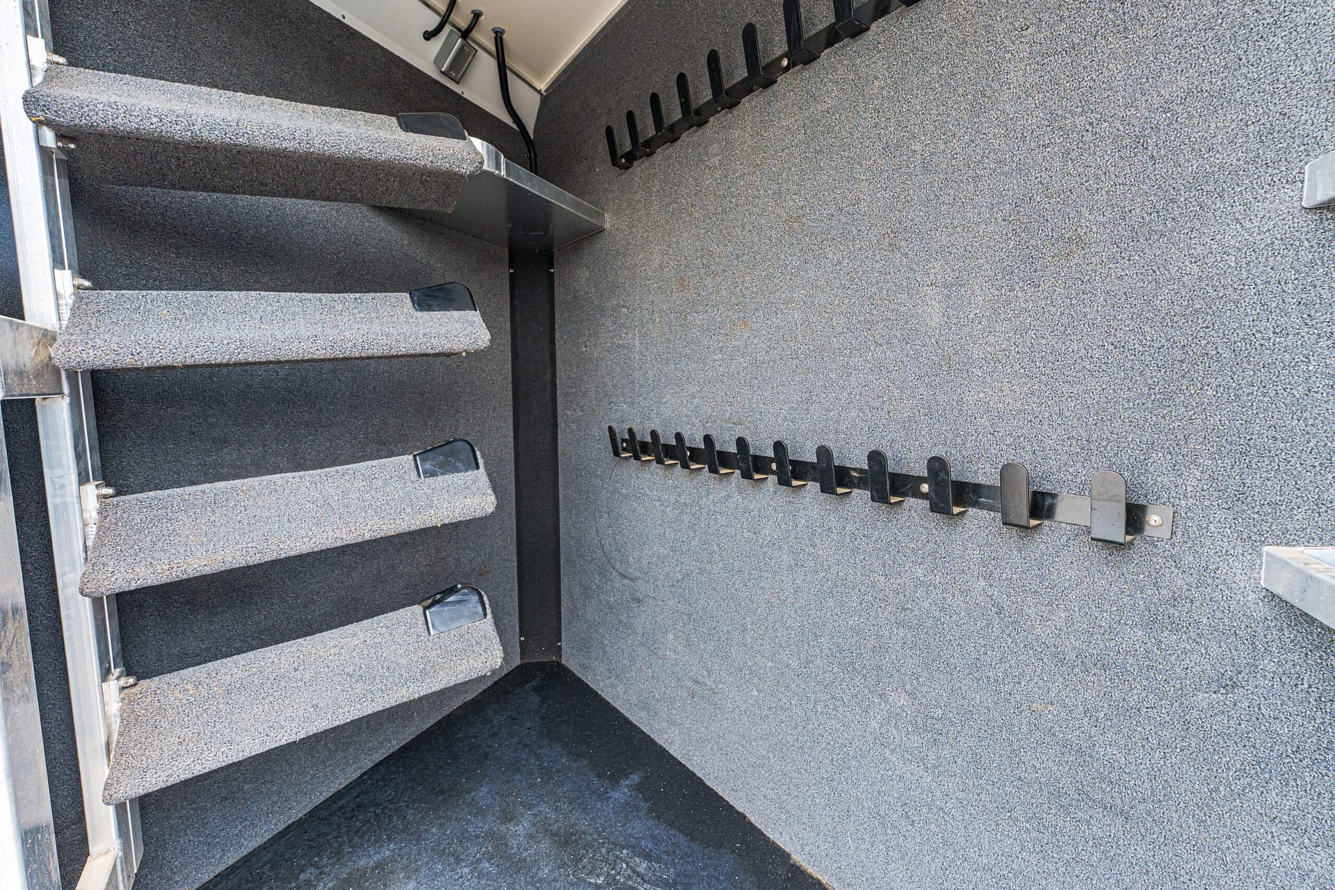 Interior of a carpeted metal staircase descending inside a gray industrial trailer; wall has a black modular tool rack and metal braces visible on the left.