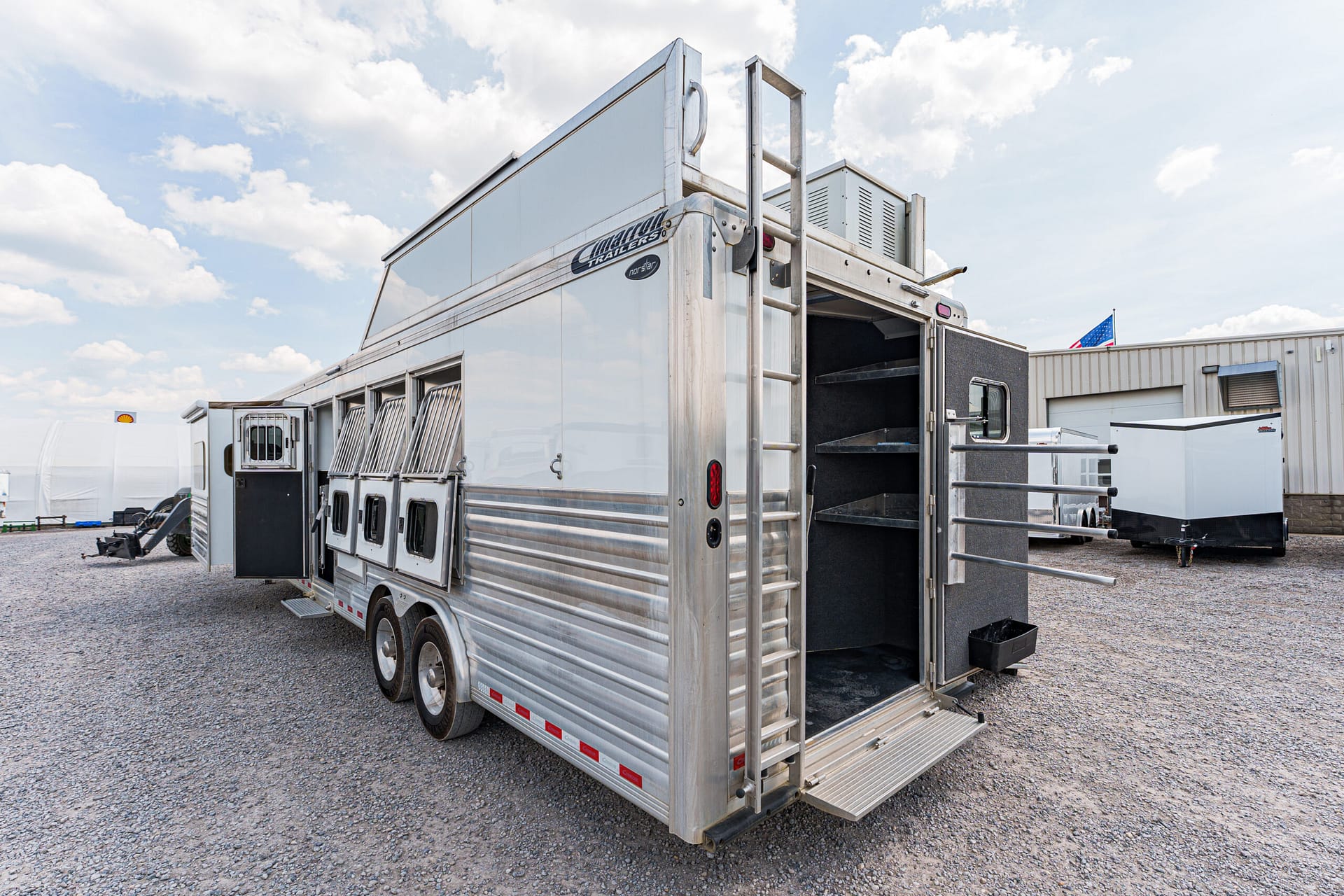 Aluminum livestock trailer with an open rear ramp, parked on a gravel lot under a blue sky with clouds.