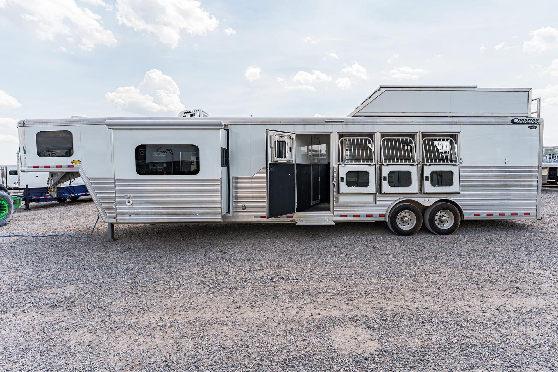Silver aluminum horse trailer with open side door and barred stall windows on a gravel lot under a blue, cloudy sky.