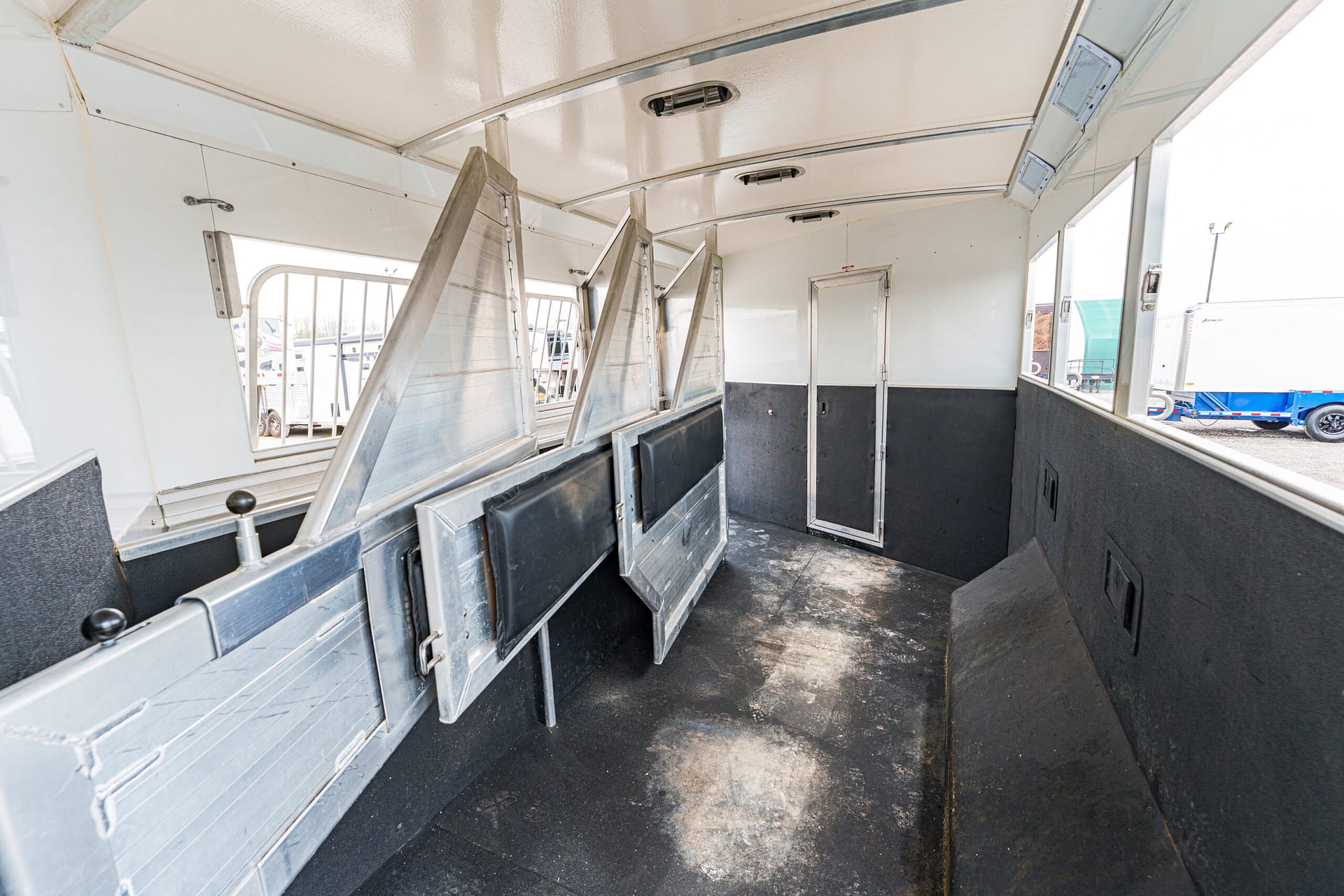 Empty transport trailer interior with angled metal stalls/dividers along the sides and a dark textured floor; light enters through side openings.