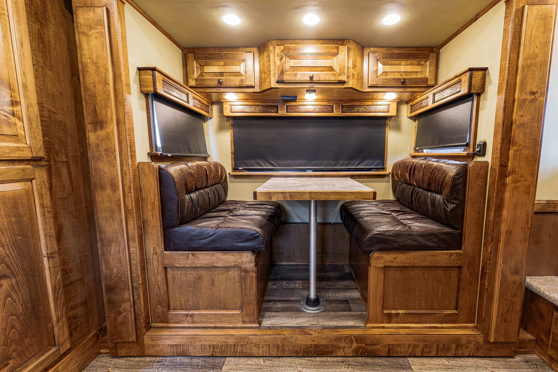 Wood-paneled RV dining area with booth seating, a small central table, and overhead storage cabinets above the windows.