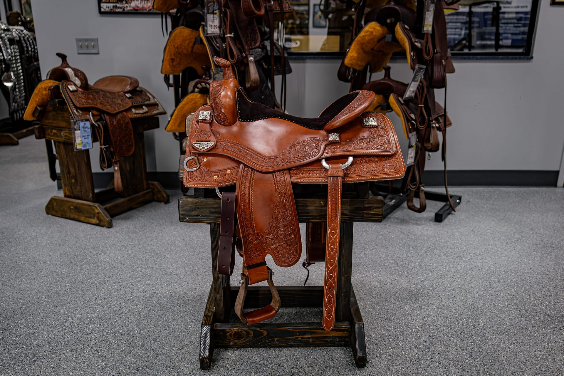 Brown leather western saddle on a wooden rack in a tack shop, with floral tooling and silver hardware visible