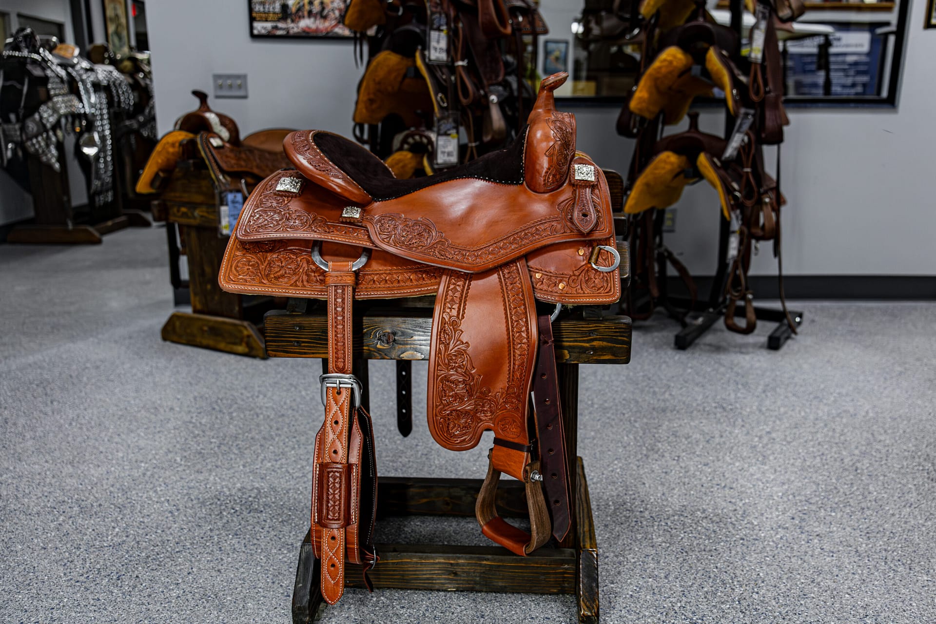 Ornately tooled brown leather western saddle on display in a tack shop, with other saddles in the background.