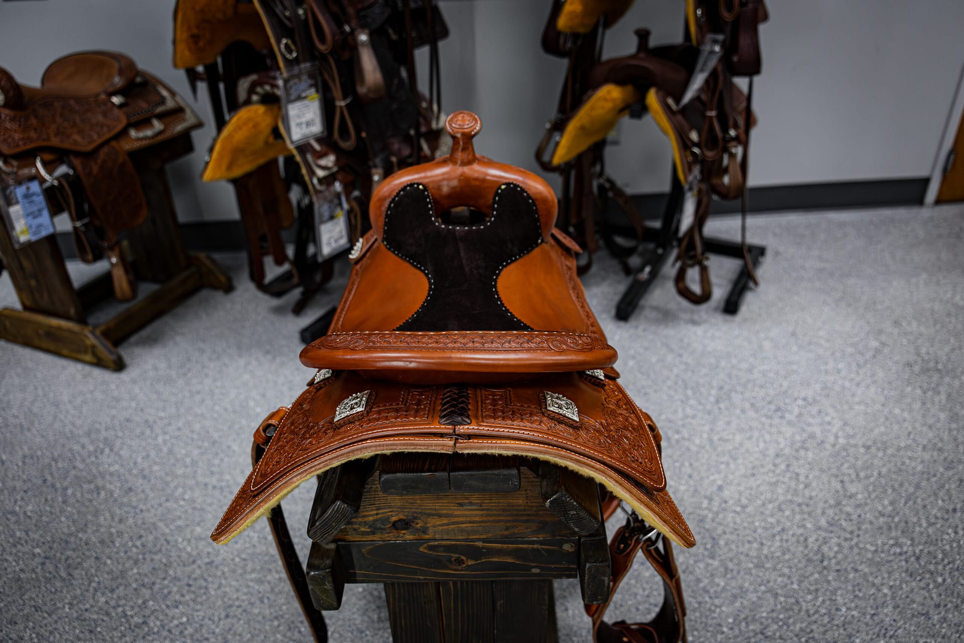 Close-up of a brown leather western saddle with black suede seat and silver stud trim, on a rack with other saddles in the background