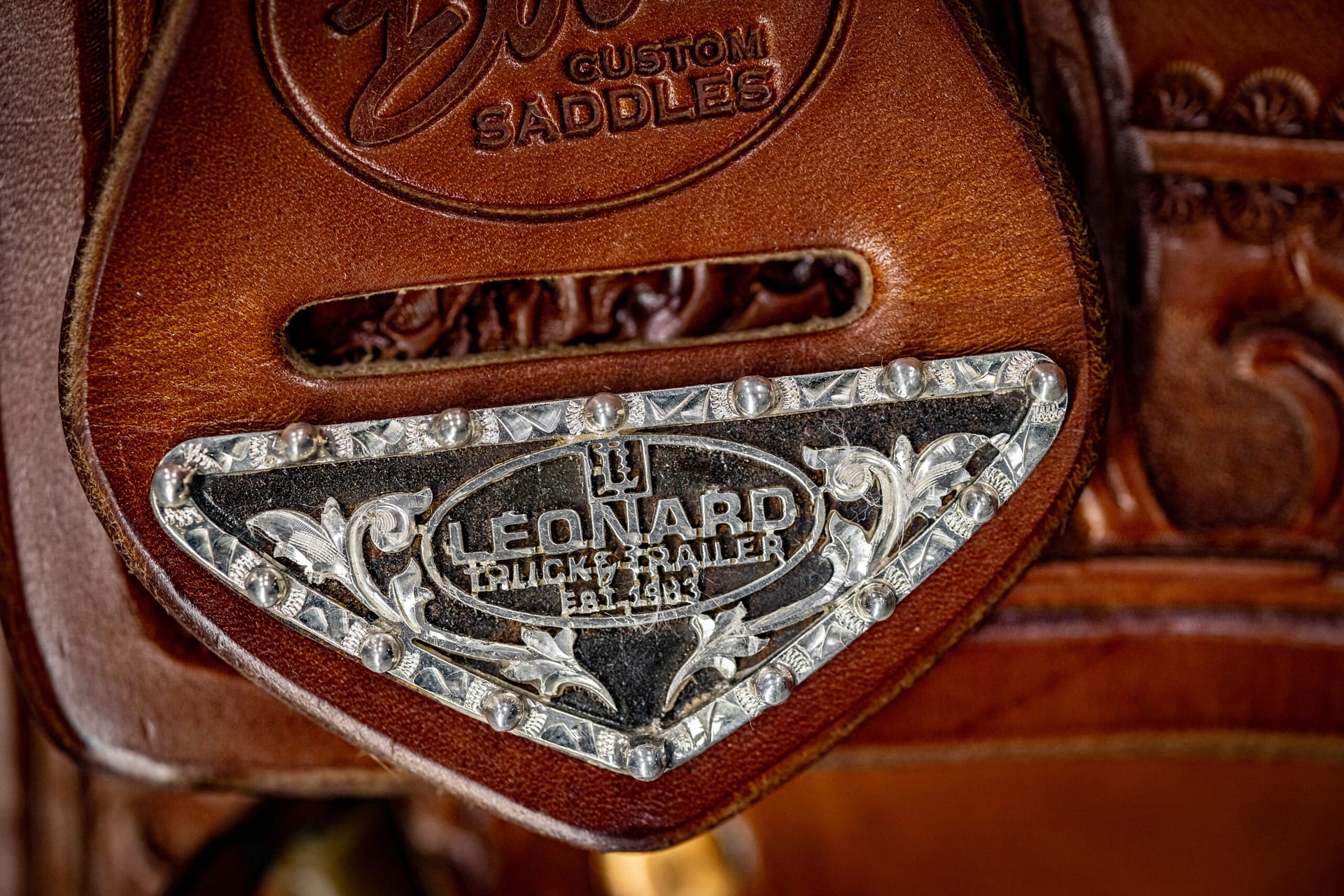 Close-up of a brown leather western saddle with an ornate silver plate and metal studs, showing detailed engraving and leatherwork.