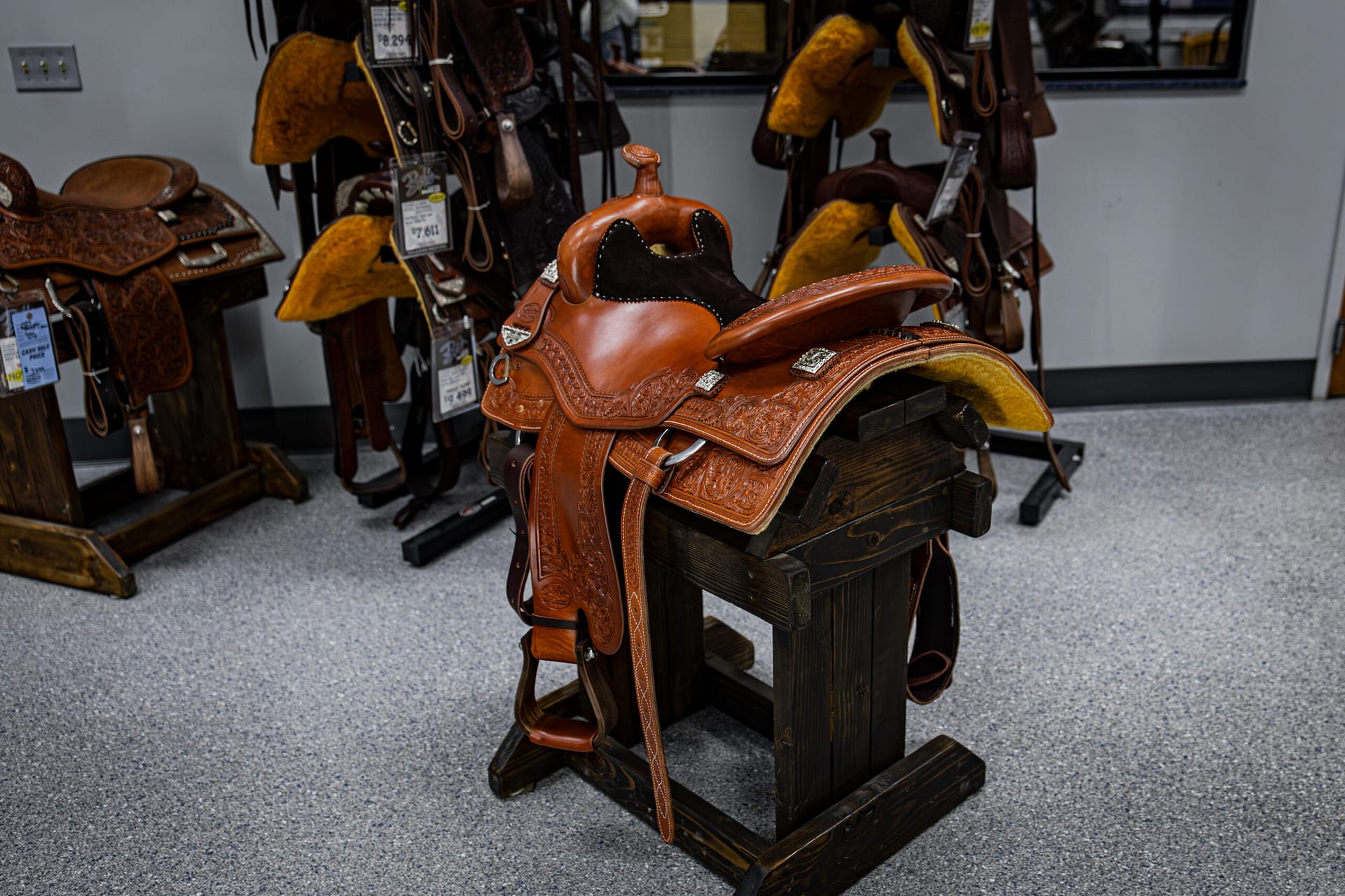 Ornate brown leather Western saddle on a wooden stand in a tack shop, with other saddles in the background.
