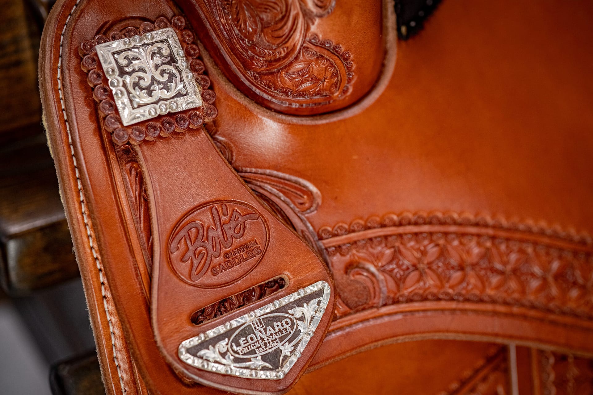 Close-up of a brown leather Western saddle with ornate silver conchos and carved floral tooling details