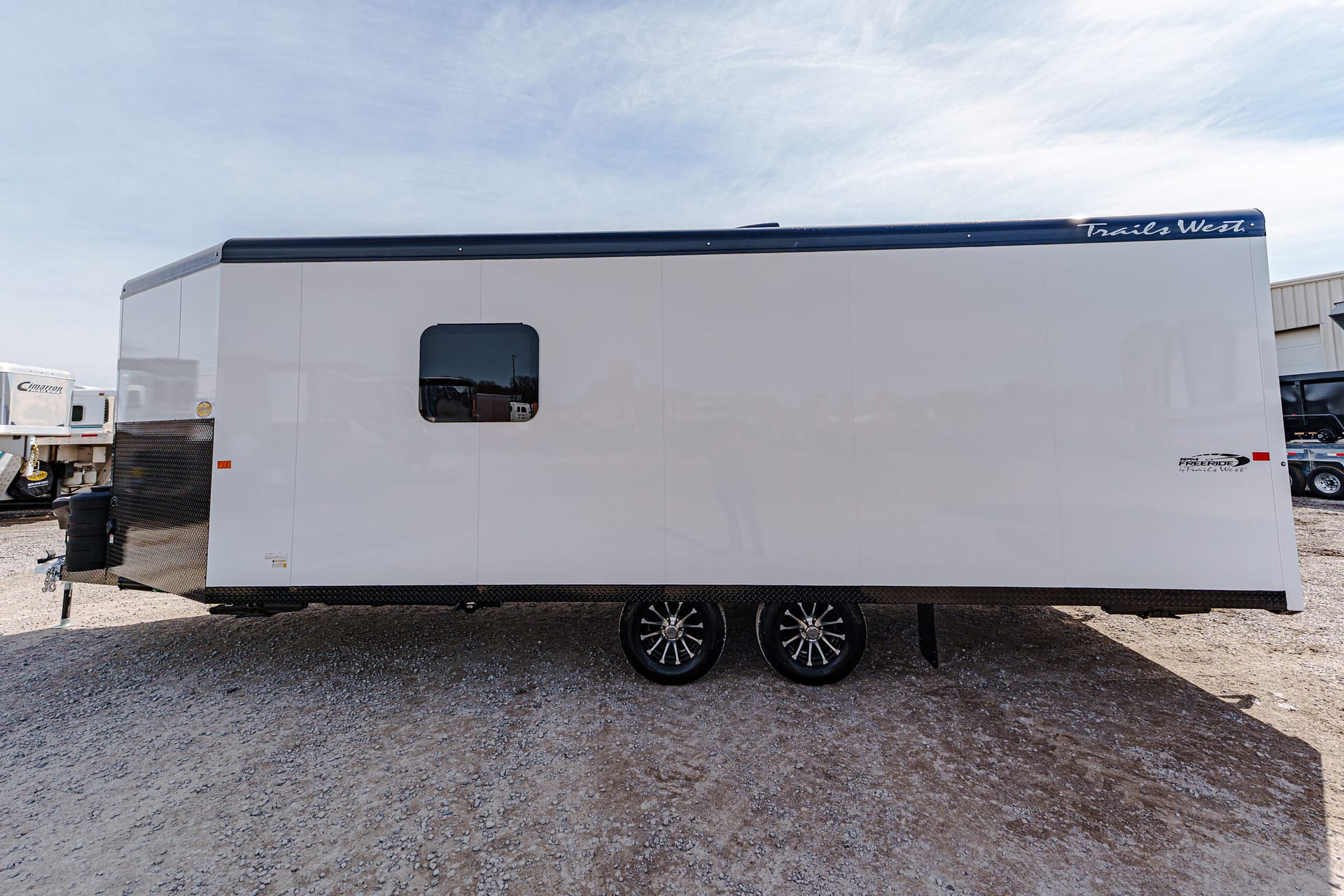White enclosed trailer with a small side window, black top trim, dual wheels, and Trails West logo on the top edge.