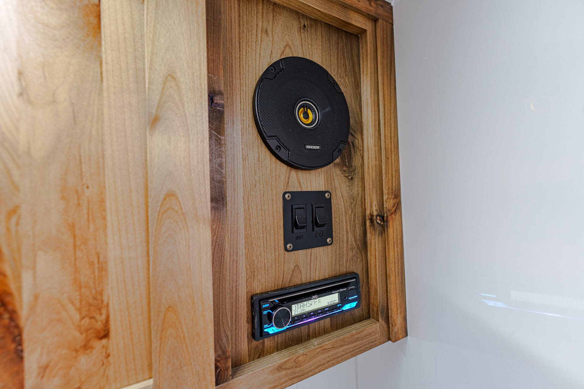 Wooden wall with a circular black speaker and a car stereo below, mounted in a wooden cabinet.