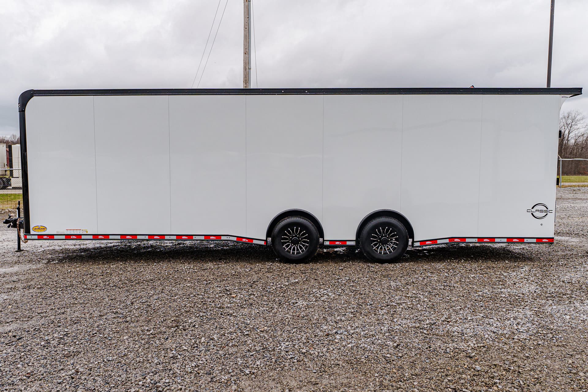 Long white enclosed cargo trailer with dual rear axles parked on gravel under a gray, cloudy sky.