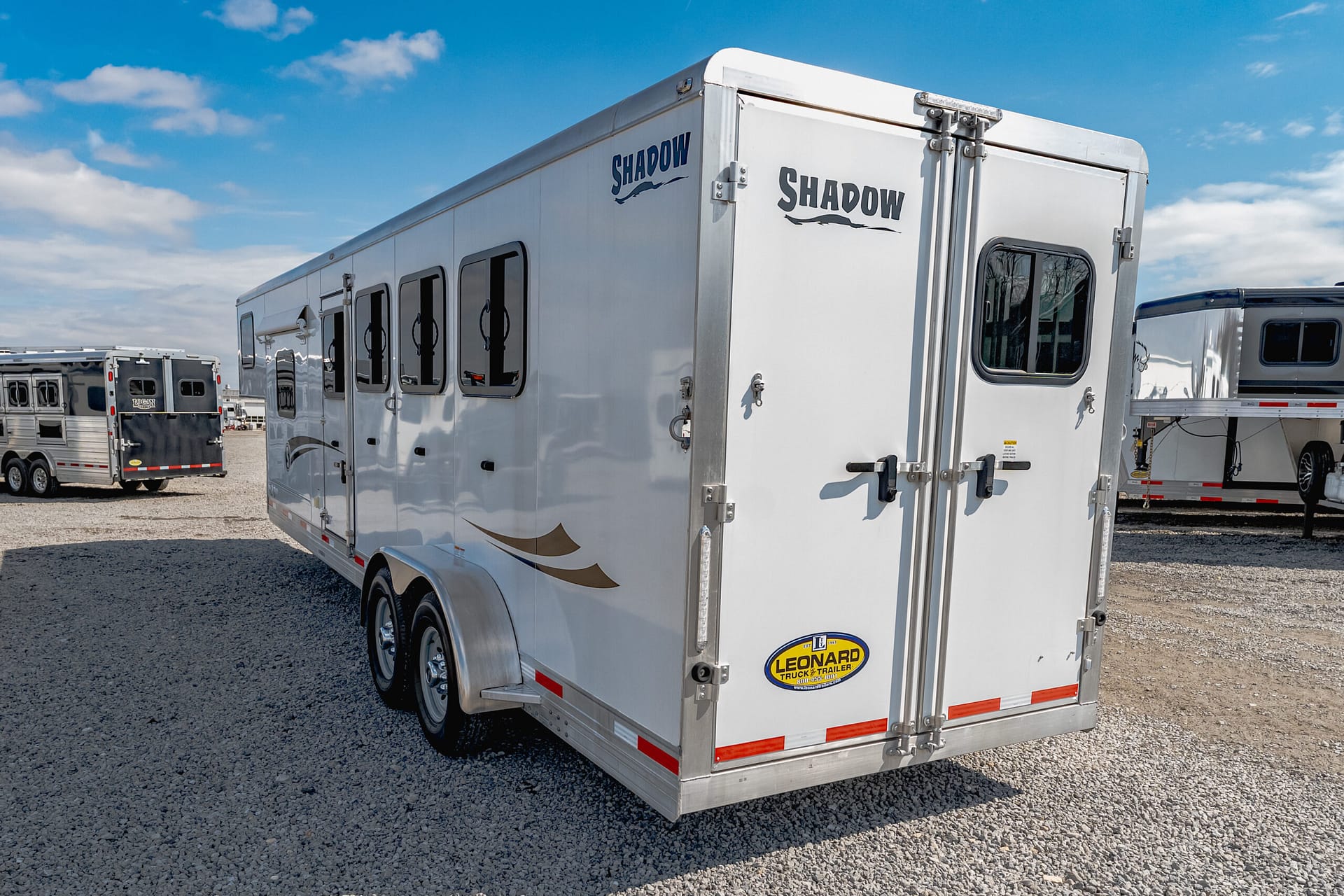 Rear-and-side view of a large white Shadow-branded horse trailer parked on a gravel lot under a blue sky with scattered clouds.
