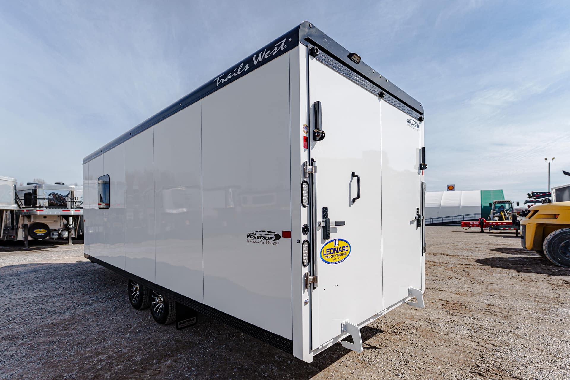 Rear view of a tall white enclosed trailer in a gravel lot with other trailers in the background on a sunny day.