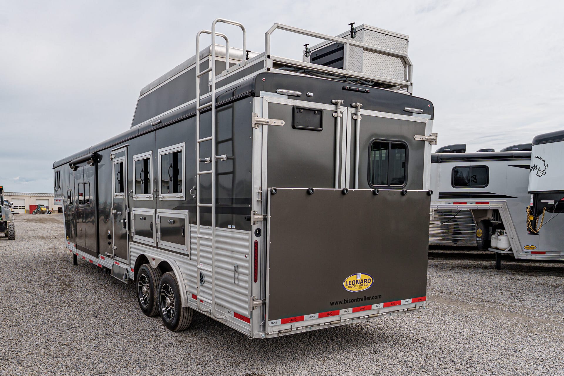 Rear and side view of a gray utility trailer with ladders, metal trim, and storage compartments on a gravel lot.