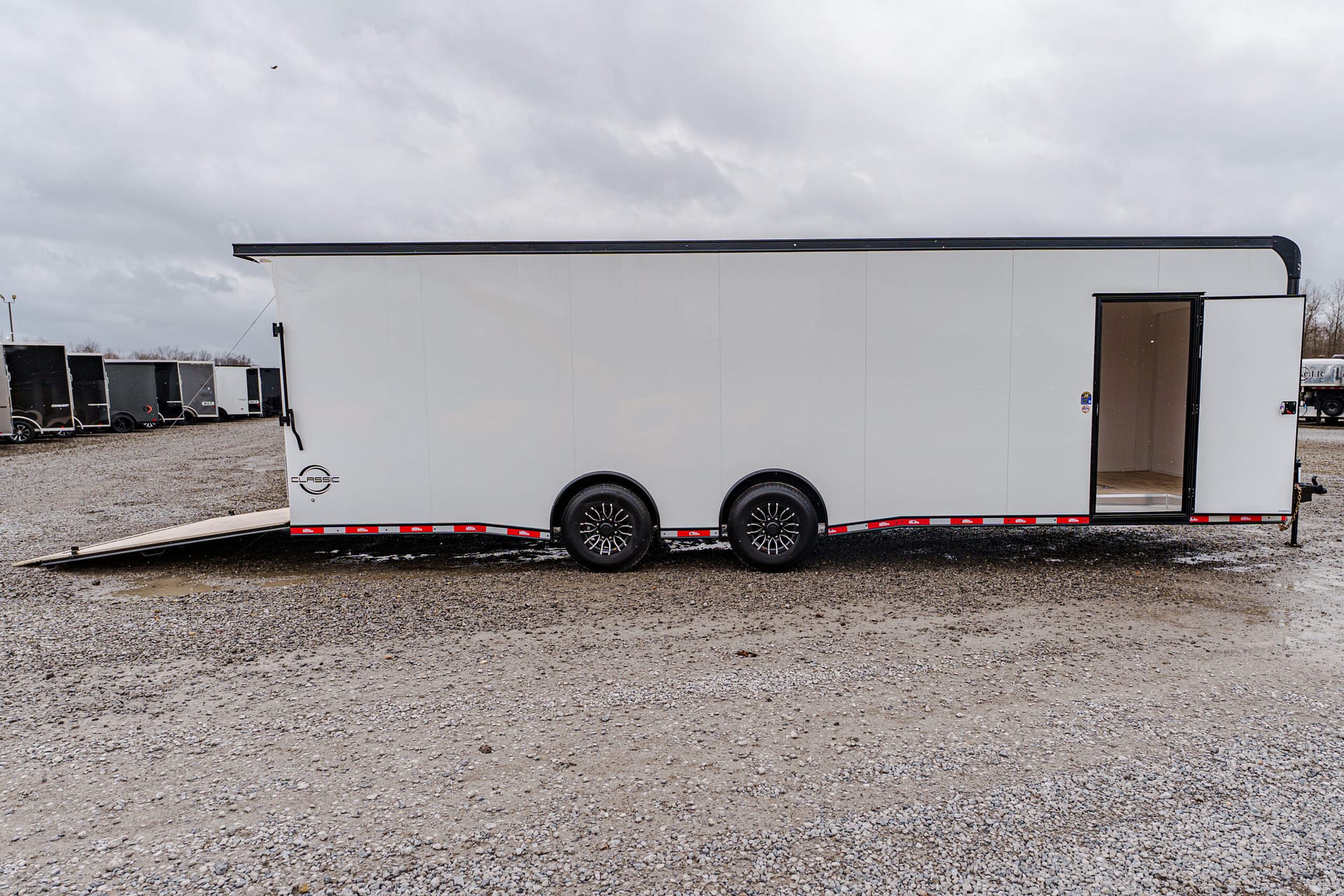 White cargo trailer with ramp lowered for loading on a gravel lot under a cloudy sky, with other trailers in the background.