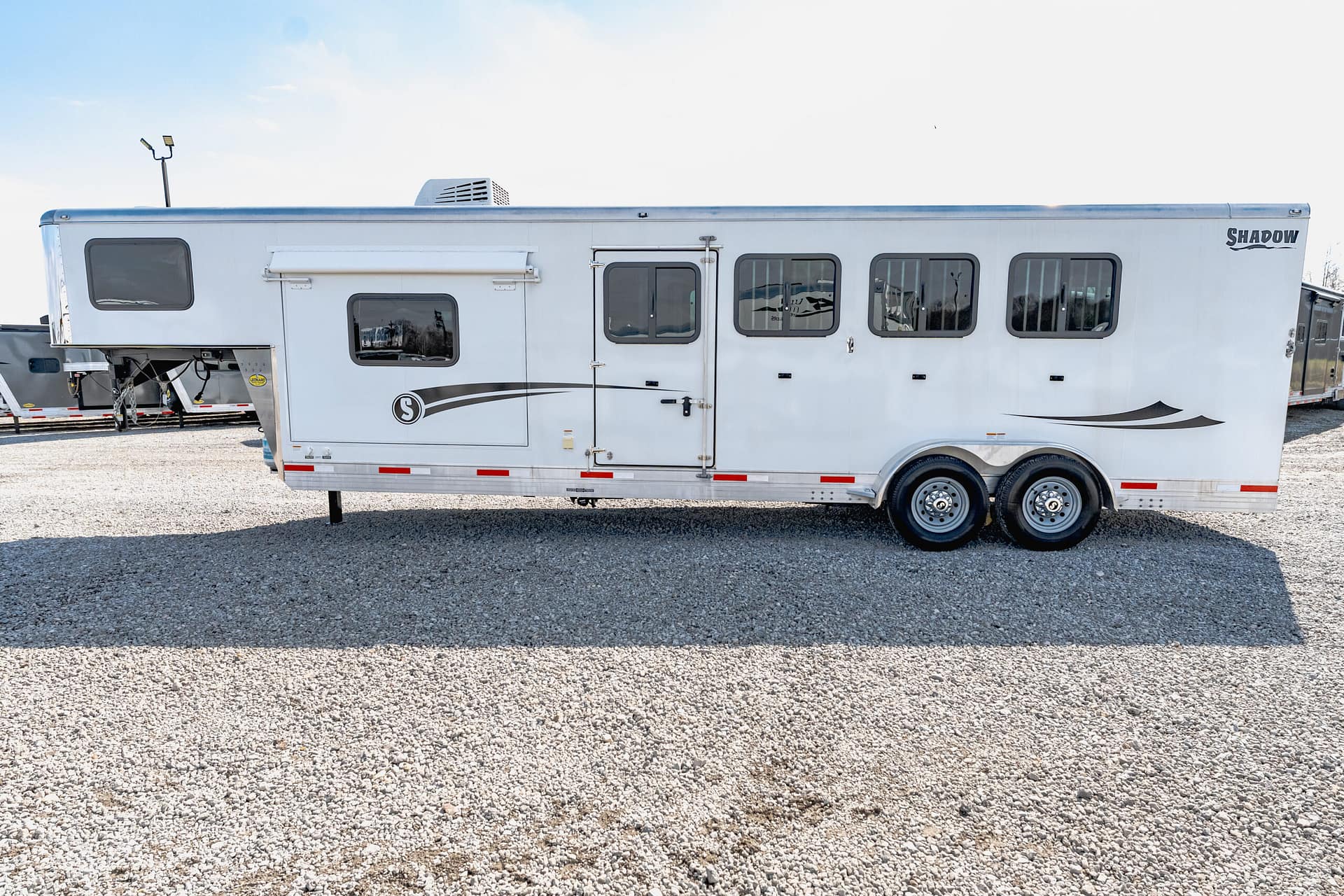 Long white travel trailer with multiple windows and a side entry door, parked on a gravel lot; 'Shadow' branding on the rear.