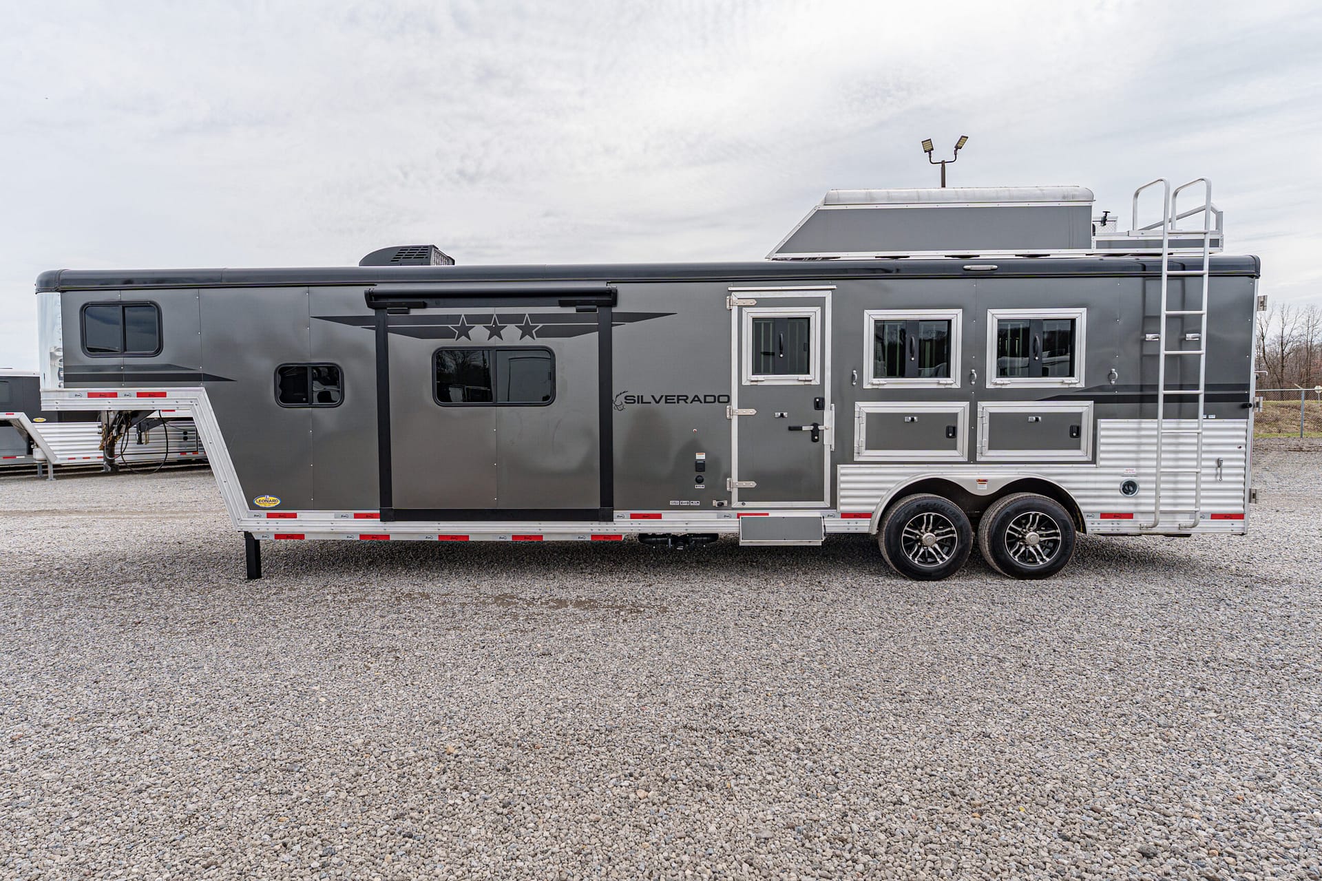 Gray Silverado travel trailer with white-framed windows, storage bays, and a ladder on a gravel lot under a cloudy sky.