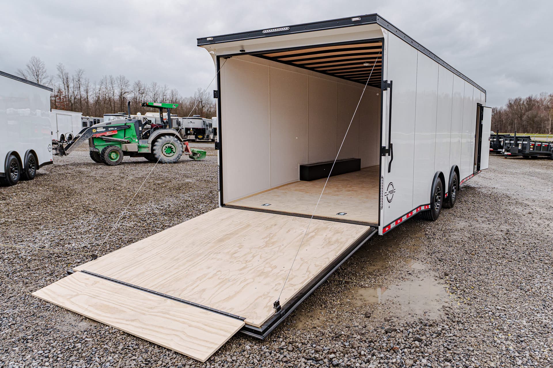 White enclosed trailer with its rear ramp lowered onto a gravel lot, empty interior and plywood floor; equipment and a green tractor visible in the background.
