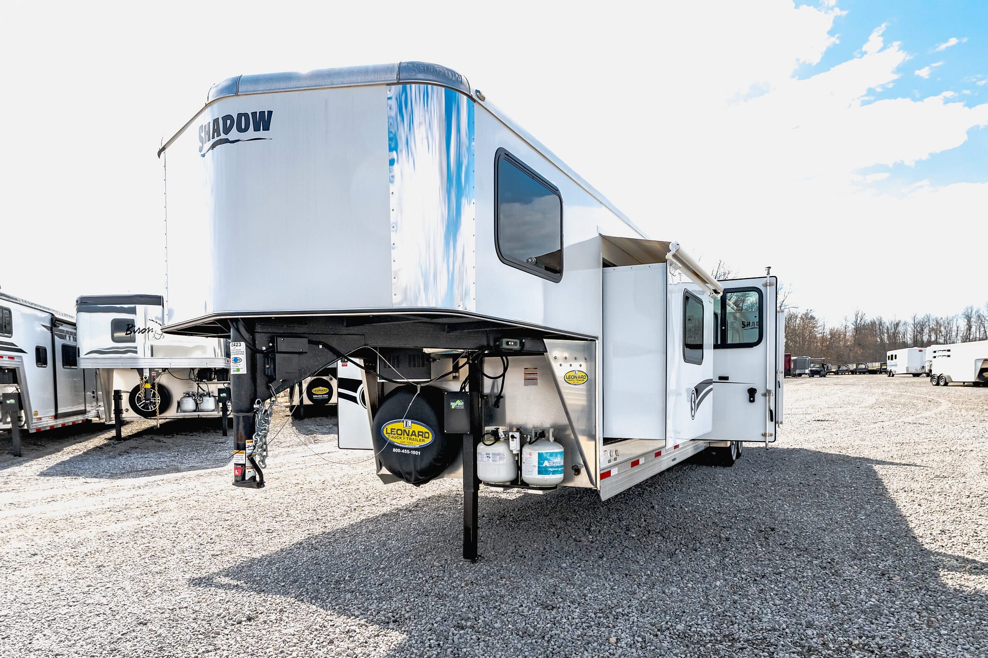 Large white fifth-wheel RV trailer with 'SHADOW' branding parked on a gravel lot, connected to a truck, with other trailers in the background.